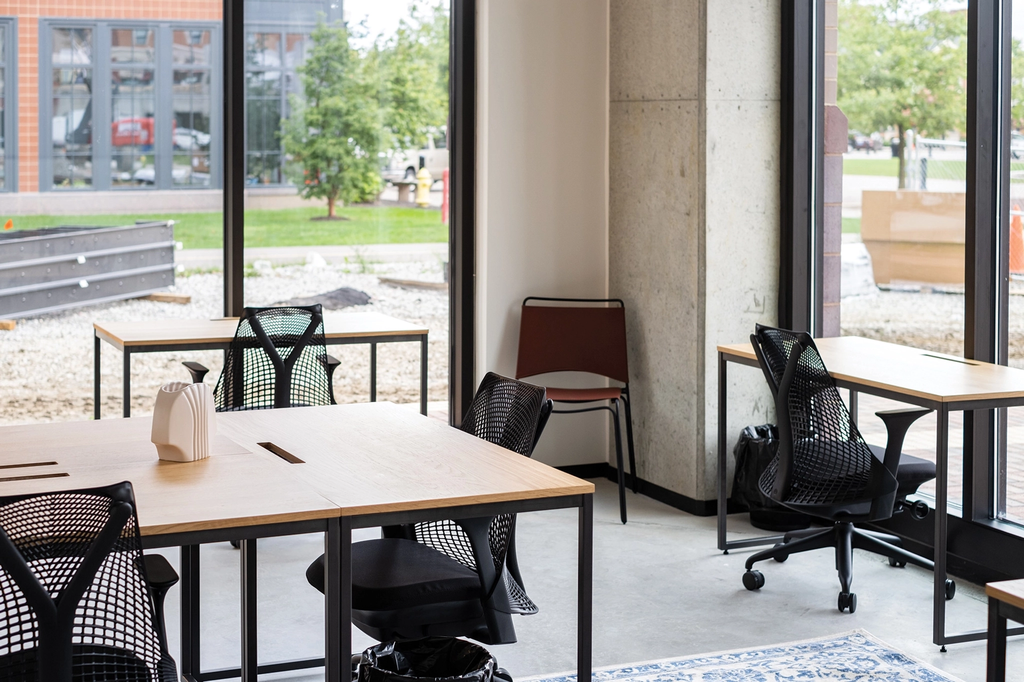 An empty, modern workspace in El Segundo features wooden desks, black mesh chairs, large windows, and a serene view of the trees outside.