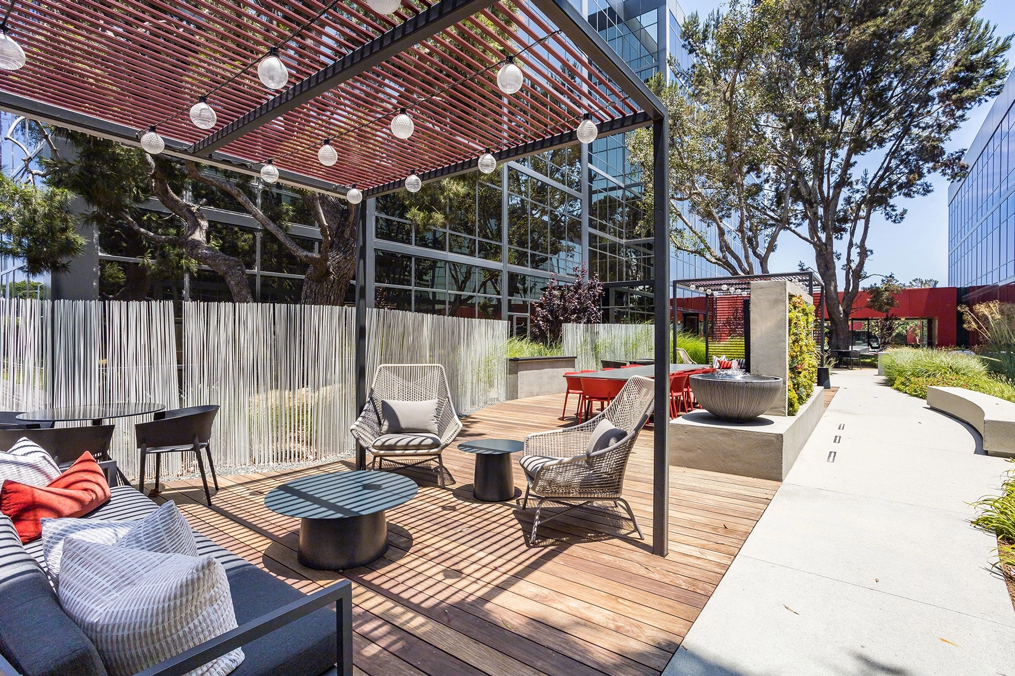 The outdoor patio area in El Segundo features modern seating, tables, and string lights. This inviting space is perfect for a coworking session or an inspiring meeting room alternative, surrounded by trees and adjacent to a sleek glass building.