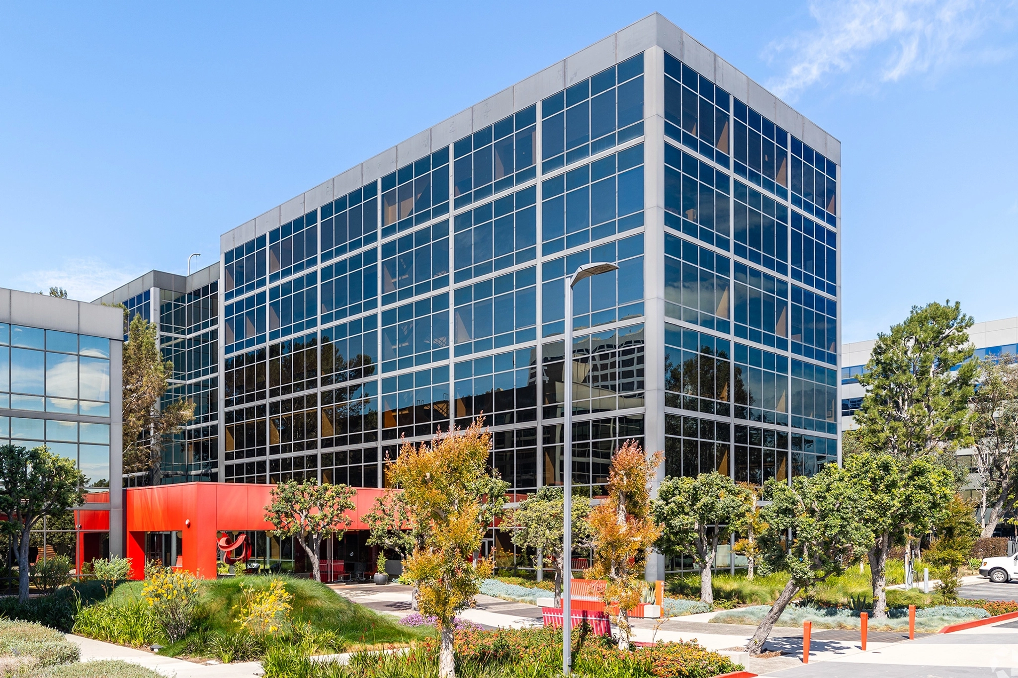 Un moderno edificio de oficinas de vidrio en El Segundo cuenta con varios pisos rodeados de árboles y vegetación bajo un cielo azul claro. En el interior, los espacios de reunión y coworking de última generación están dirigidos a profesionales que buscan un entorno de trabajo dinámico e inspirador.