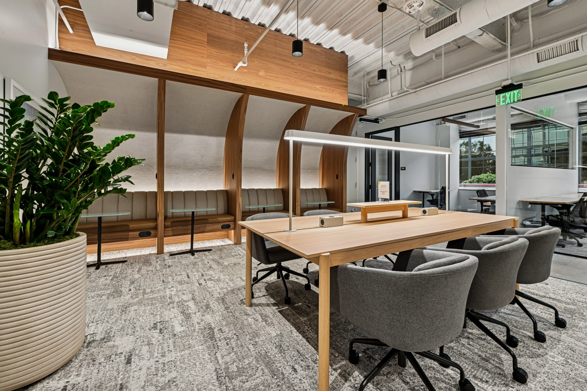 Moderner Coworking-Bürobereich in El Segundo mit einem langen Holzschreibtisch, grauen Stühlen, integrierten Sitzgelegenheiten, einer großen Topfpflanze und einem verglasten Konferenzraum im Hintergrund.