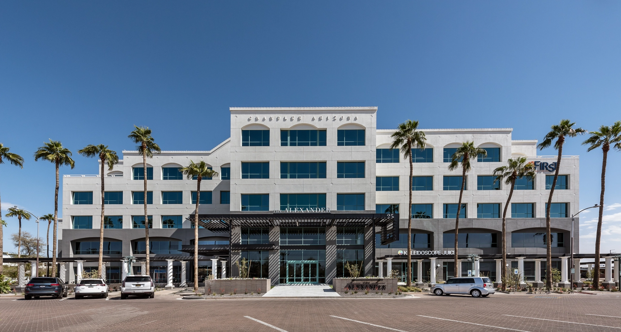 Front view of The Alexander building with a glass facade, metal canopy, and surrounding greenery. Address number 25 is displayed.