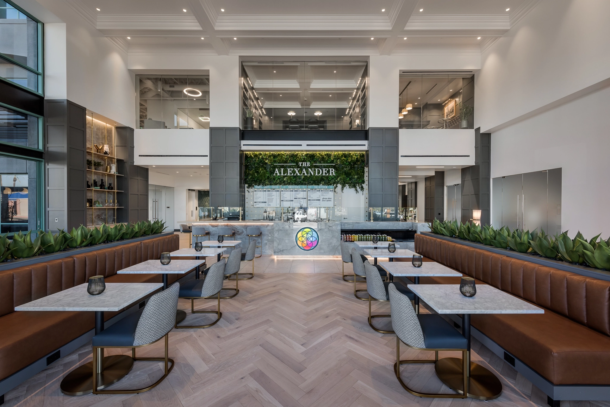 Spacious modern café interior with marble tables, cushioned seating, and green plants. A counter with a sign "The Alexander" and a menu is in the background, complemented by high ceilings and bright lighting.