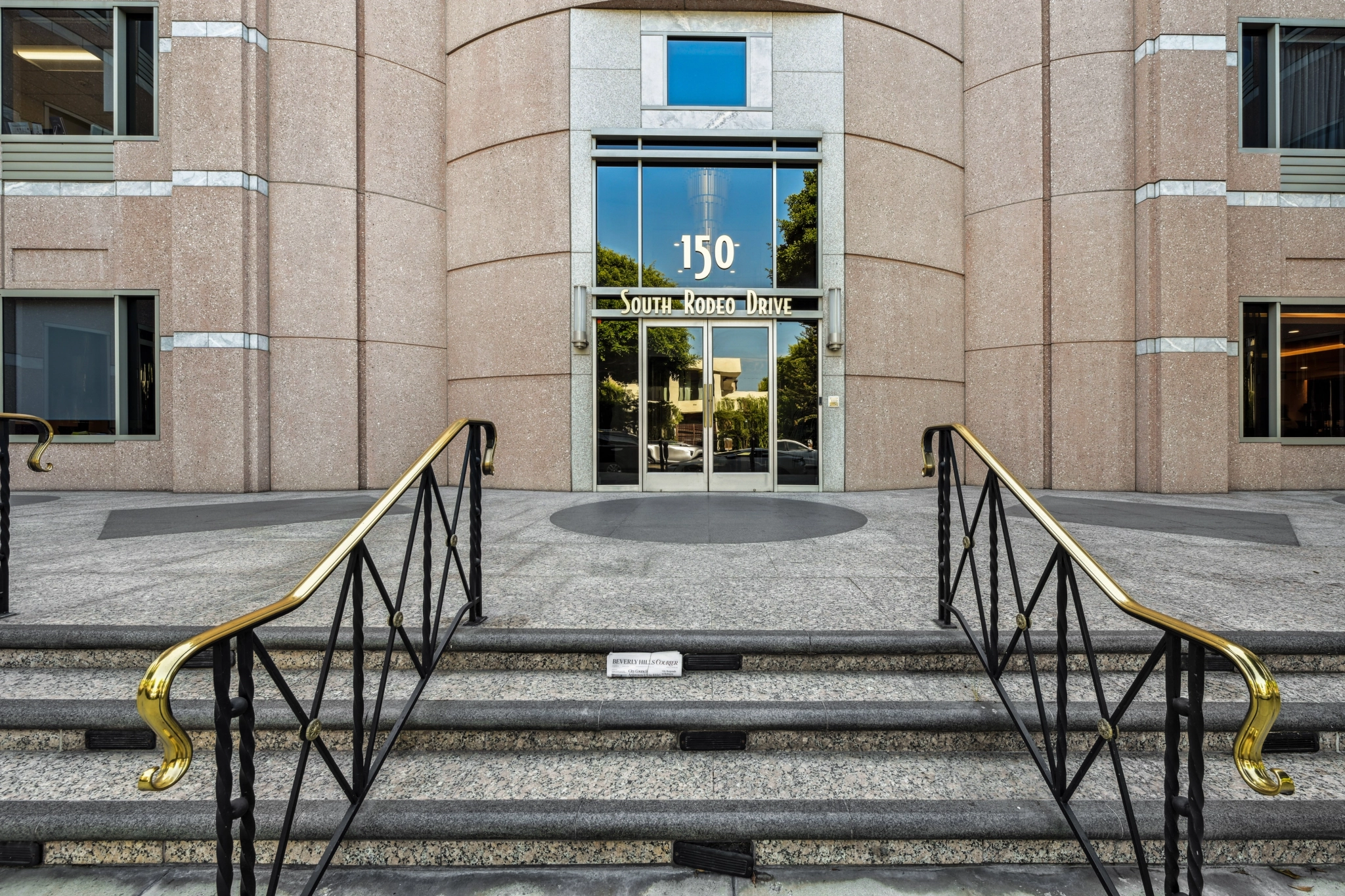 Entrée d'un immeuble de bureaux moderne situé au 150 South Rodeo Drive à Beverly Hills, avec des marches en granit, des rampes noires et dorées, et des portes en verre reflétant les arbres et les espaces de coworking voisins.