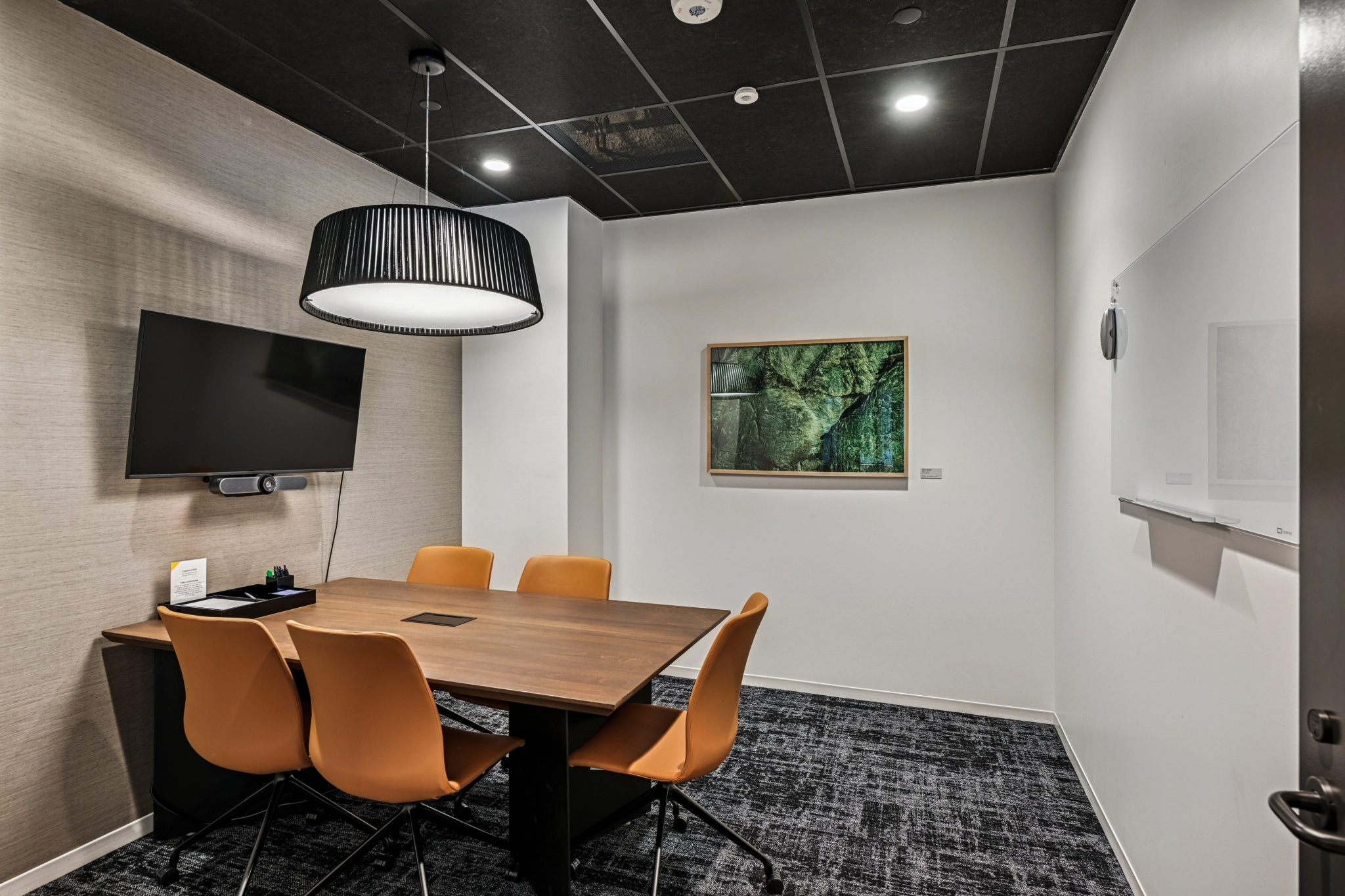 Espace de travail moderne de Beverly Hills comprenant une petite salle de conférence avec une table en bois, quatre chaises orange, une télévision murale, un tableau blanc et des œuvres d'art forestières encadrées - idéal pour des sessions de coworking ciblées.
