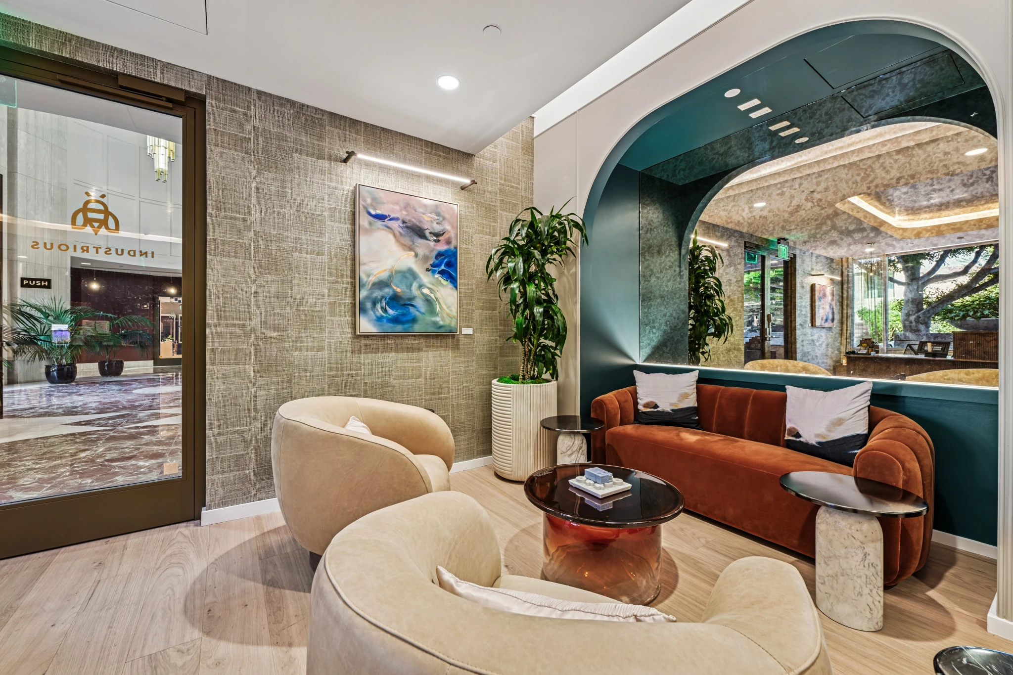Modern office lobby in Beverly Hills with beige armchairs, a rust-colored sofa, glass tables, a large plant, and abstract artwork. Glass doors and windows let in natural light, creating an inviting workspace near the meeting room.