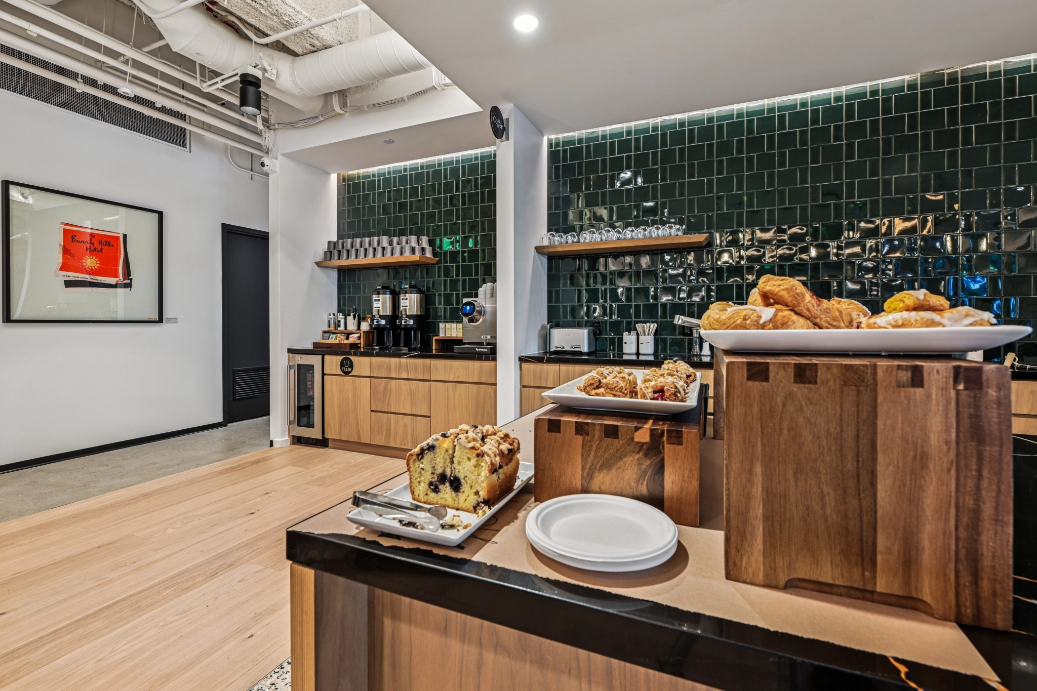 Une cuisine moderne dans un bureau de Beverly Hills, avec des viennoiseries et du pain exposés, des armoires en bois, un dosseret en carrelage vert, des machines à café et des tasses bien rangées sur des étagères.