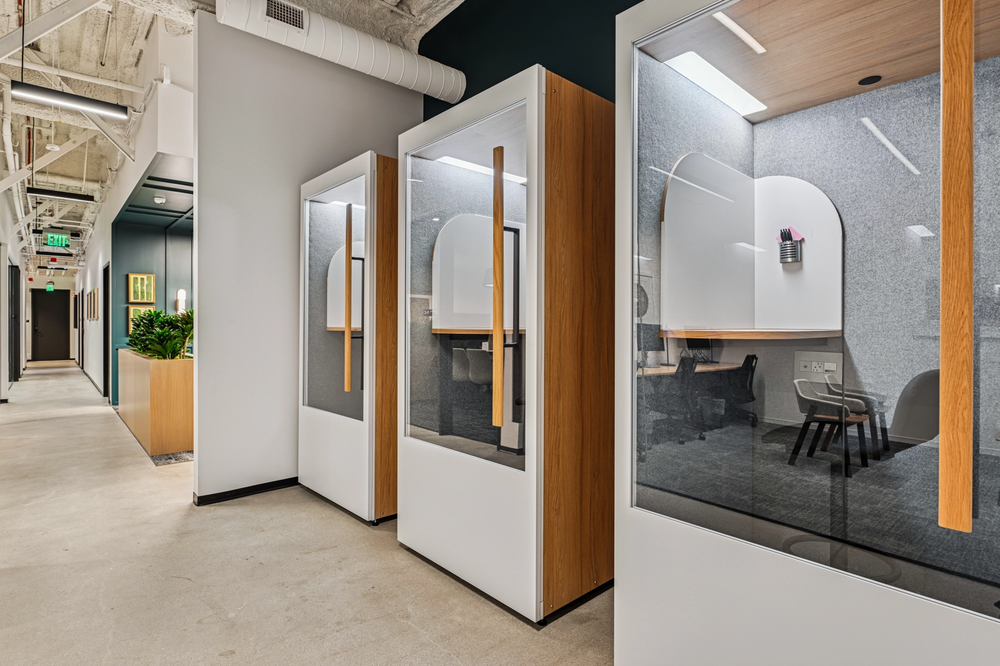 Couloir de bureau moderne avec deux cabines téléphoniques vitrées sur la gauche et une salle de réunion avec une table et des chaises sur la droite. Cet espace de travail se caractérise par des tons neutres et une décoration minimale pour un aspect épuré et contemporain.