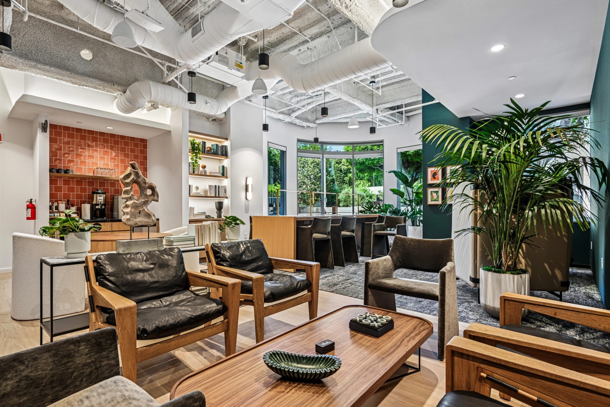 Modern coworking office lounge in Beverly Hills featuring wooden furniture, black leather chairs, large potted plants, a coffee station with red tile backsplash, bookshelves, and expansive windows flooding the space with natural light.