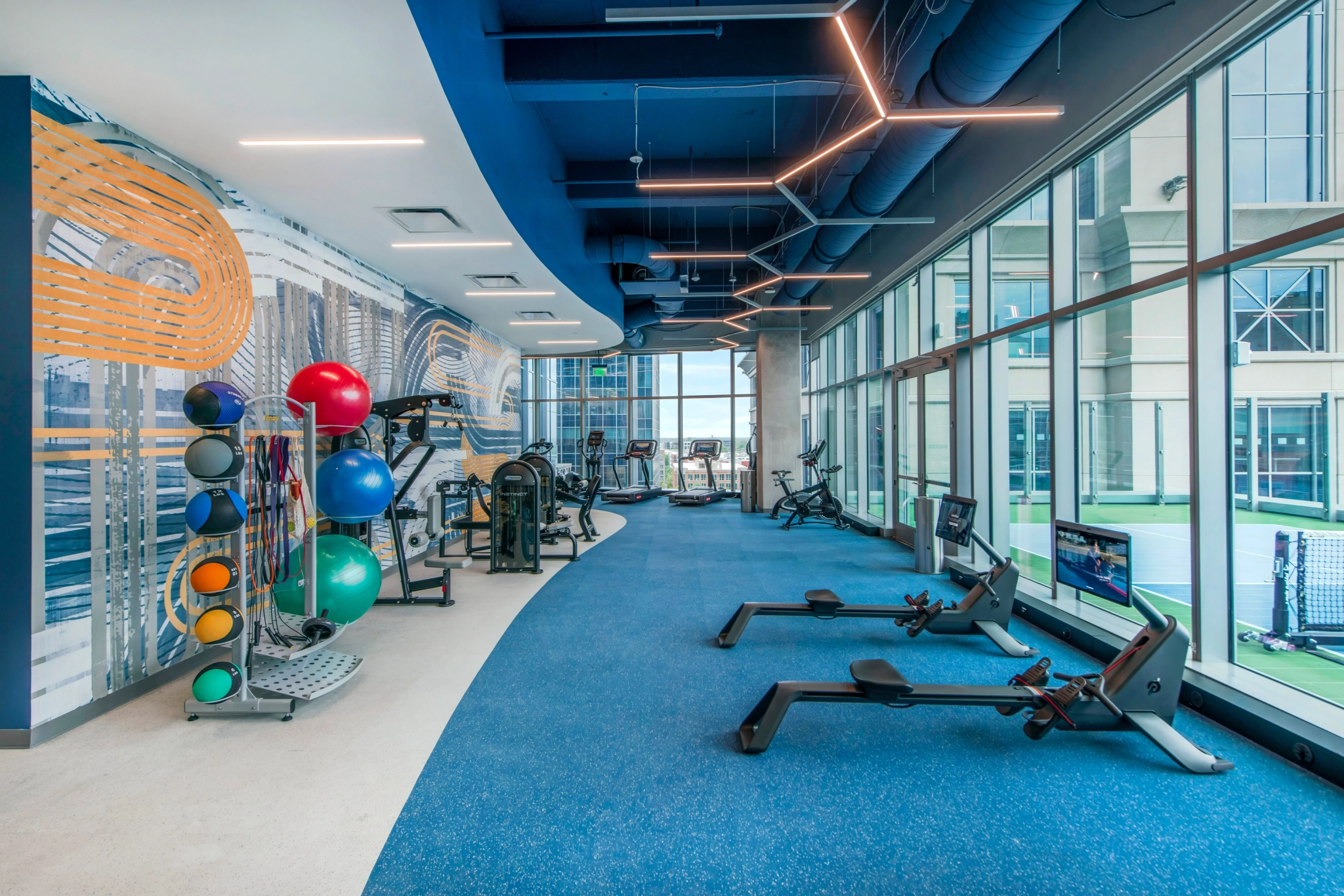 Moderno gimnasio en Charlotte con grandes ventanales, varias máquinas de fitness -incluidas bicicletas estáticas, máquinas de remo, pesas y pelotas de estabilidad- en un suelo azul, perfecto para una pausa de bienestar en la oficina o después de una reunión en la sala de conferencias.