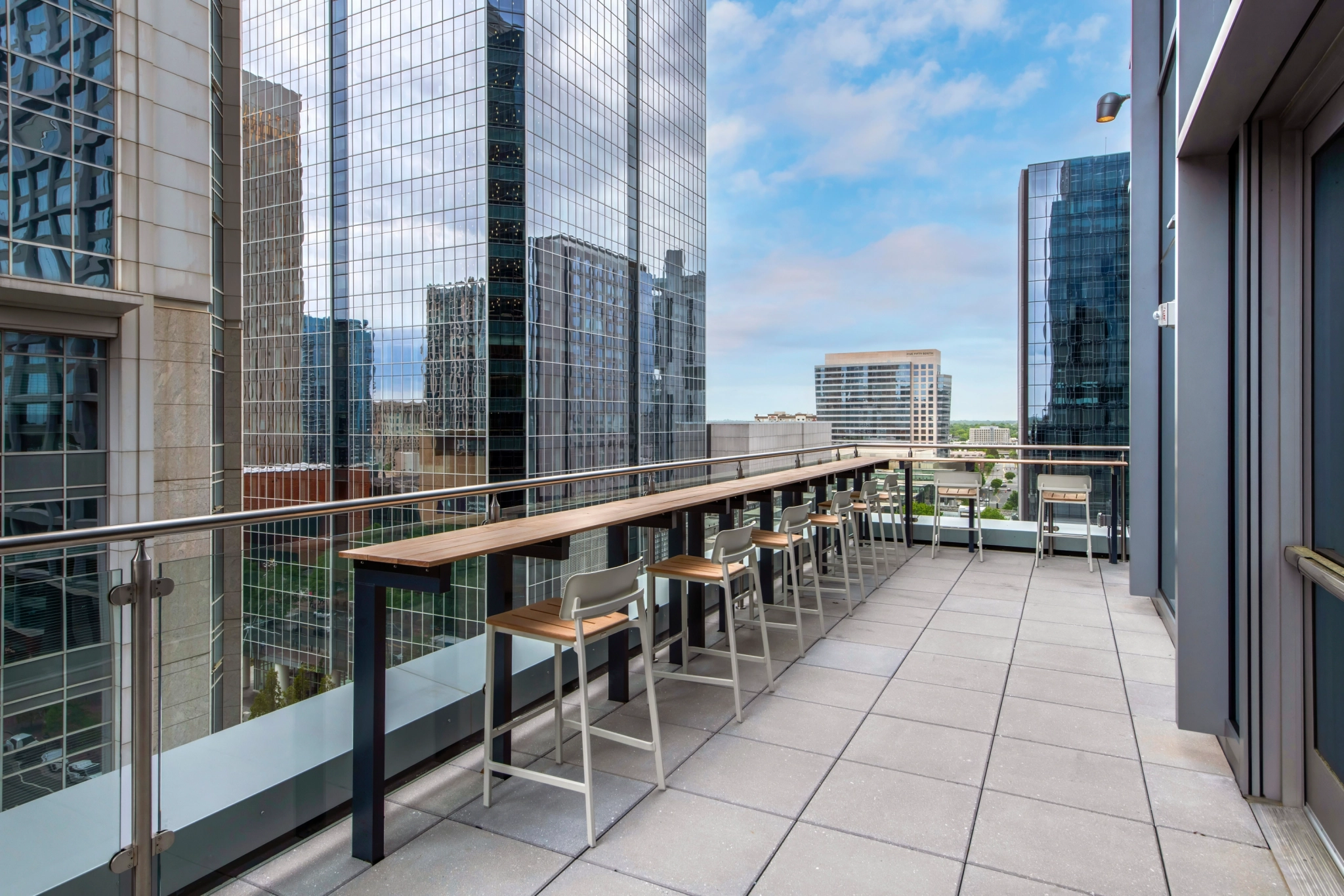 Una moderna terraza exterior con una larga mesa de bar de madera y sillas ofrece un espacio de trabajo dinámico y vistas a un paisaje urbano de altos edificios de oficinas de cristal bajo un cielo parcialmente nublado.