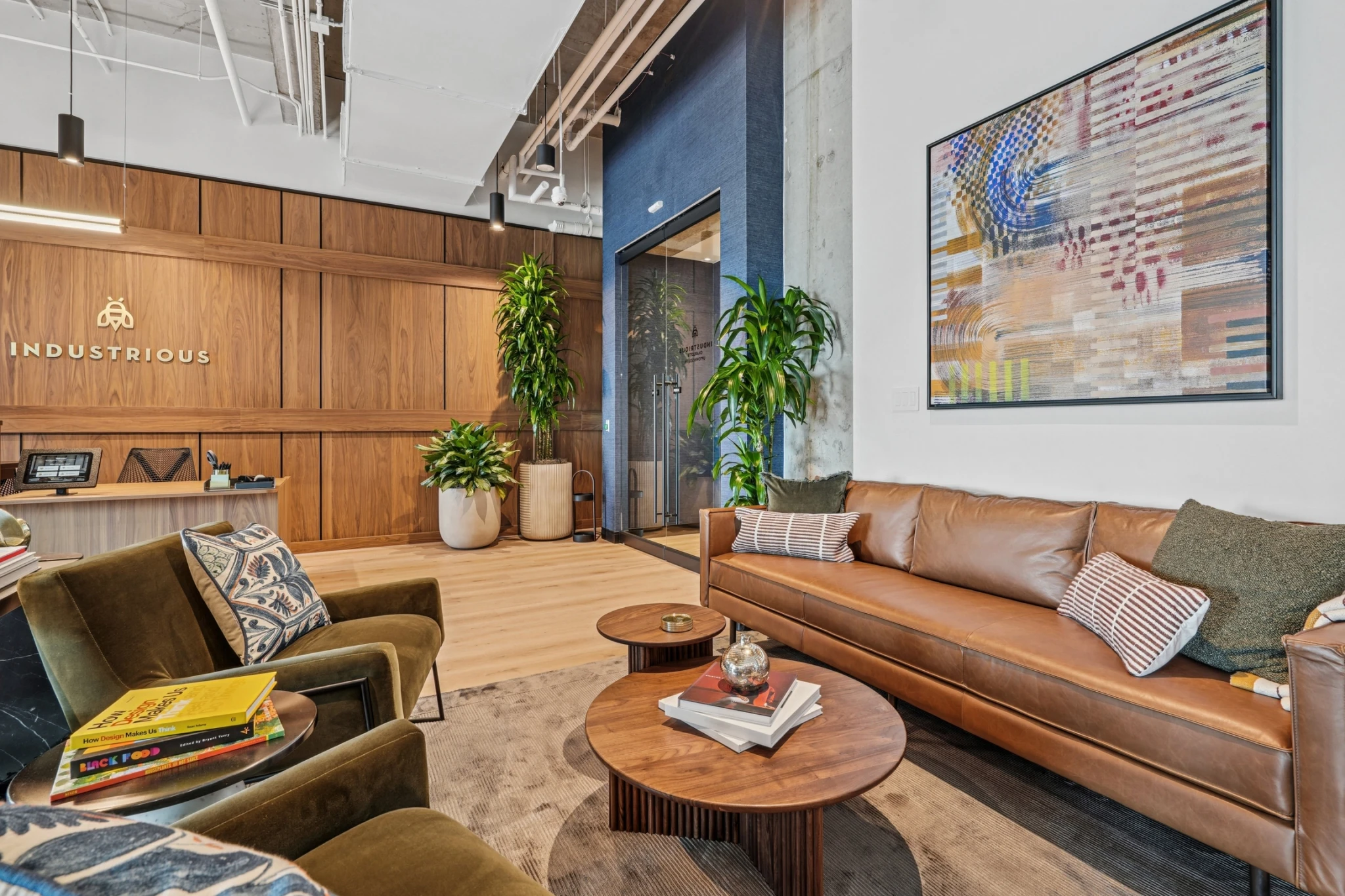 Modern coworking lounge with tan leather sofa, armchairs, wooden coffee tables with books, potted plants, abstract wall art, and wooden paneling with "Industrious" logo—perfect for informal meetings or a relaxing office break.