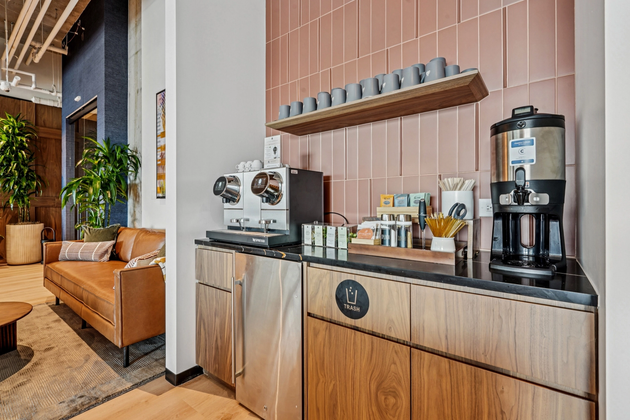 Un moderno espacio de trabajo con dos cafeteras, tazas, bolsitas de té, agitadores y un dispensador de agua sobre un mostrador de madera junto a un salón con plantas y asientos: ideal para su próxima pausa en la sala de reuniones Charlotte.