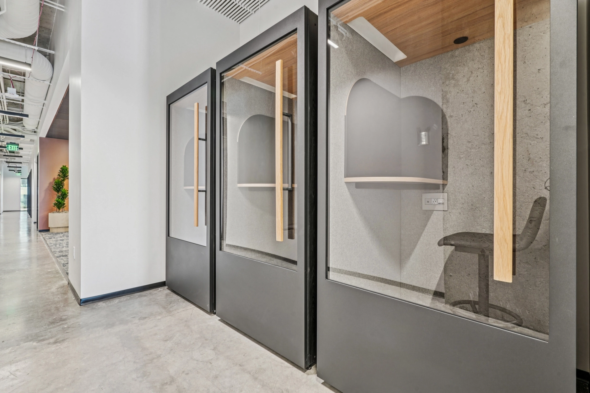 Tres modernas cabinas telefónicas de escritorio con puertas de cristal y tiradores de madera se alinean en un pasillo del espacio de trabajo de Charlotte con suelo de hormigón pulido y techo visto.