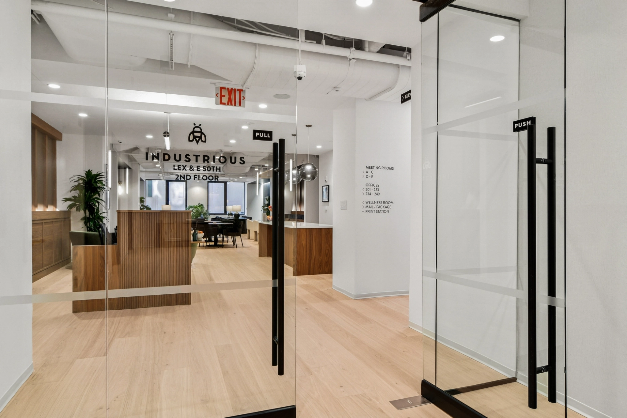 Modern office entrance with glass doors labeled "Push" and "Pull," leading to a stylish workspace reception area with wood accents and "Industrious" signage on the back wall.