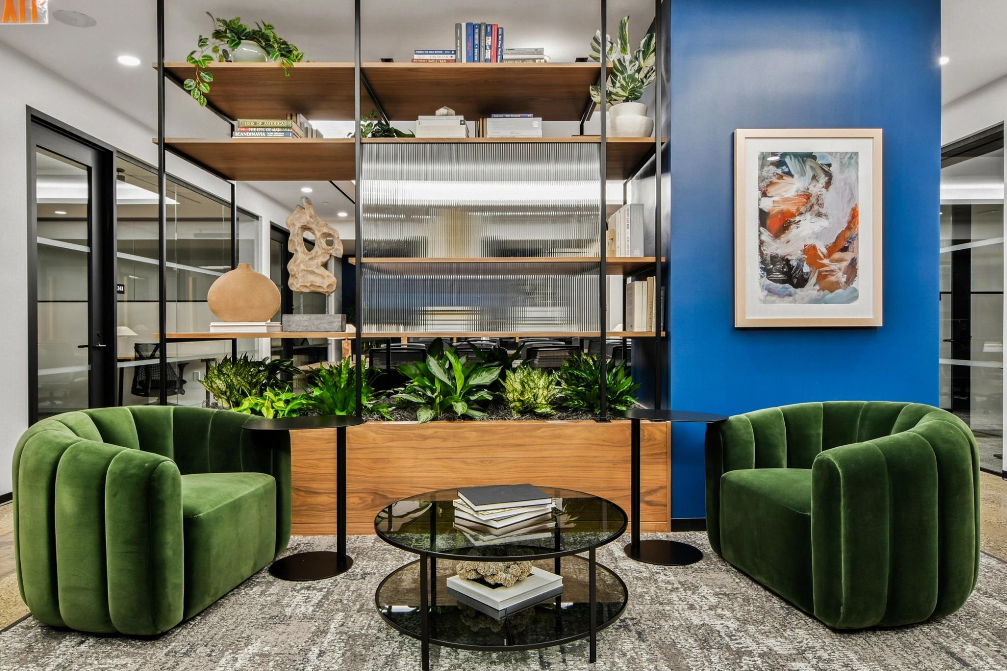 Salon de coworking moderne comprenant deux chaises en velours vert, une table basse en verre avec des livres, des plantes, un mur d'accent bleu avec des œuvres d'art, et des étagères ouvertes avec des objets décoratifs - idéal pour des réunions de bureau informelles ou pour se détendre.