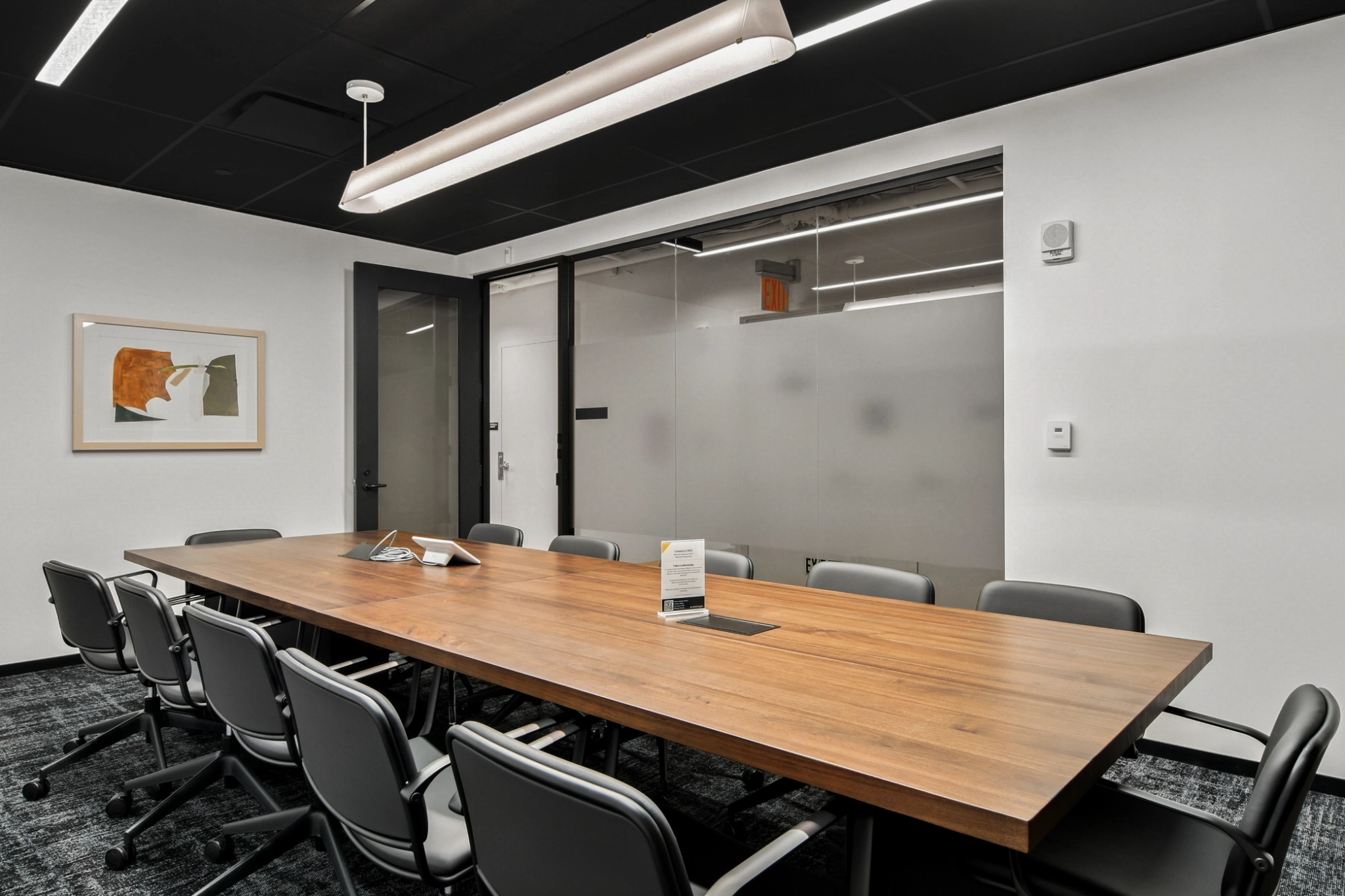 Salle de conférence moderne avec une longue table en bois, dix chaises noires, un mur en verre dépoli, des œuvres d'art encadrées et un décor minimaliste - parfait pour un espace de travail élégant ou un environnement de coworking.