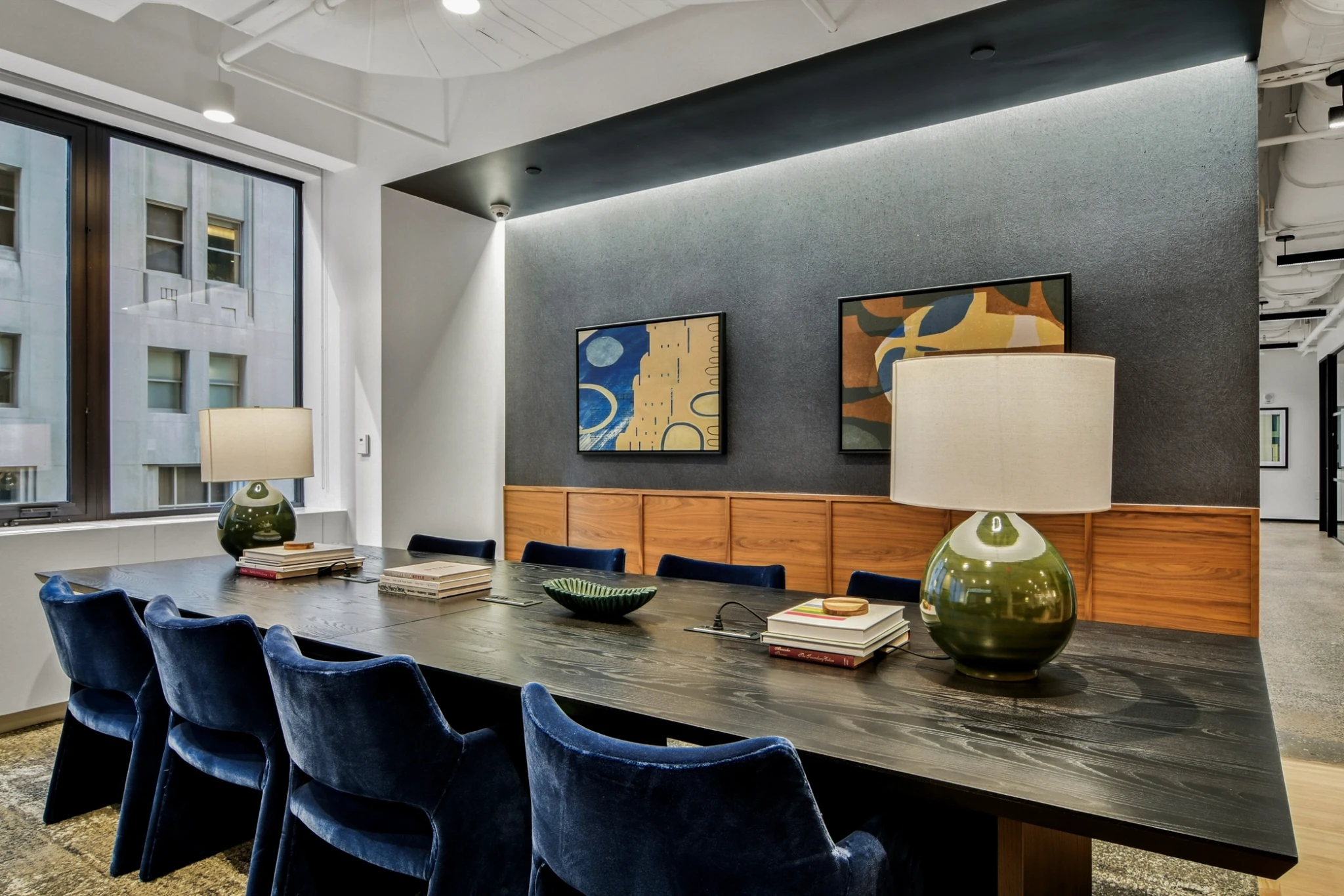 A modern meeting room with a long dark table, blue velvet chairs, two table lamps, books, and abstract art on the wall. Large windows reveal office buildings, making it an ideal workspace for collaboration.