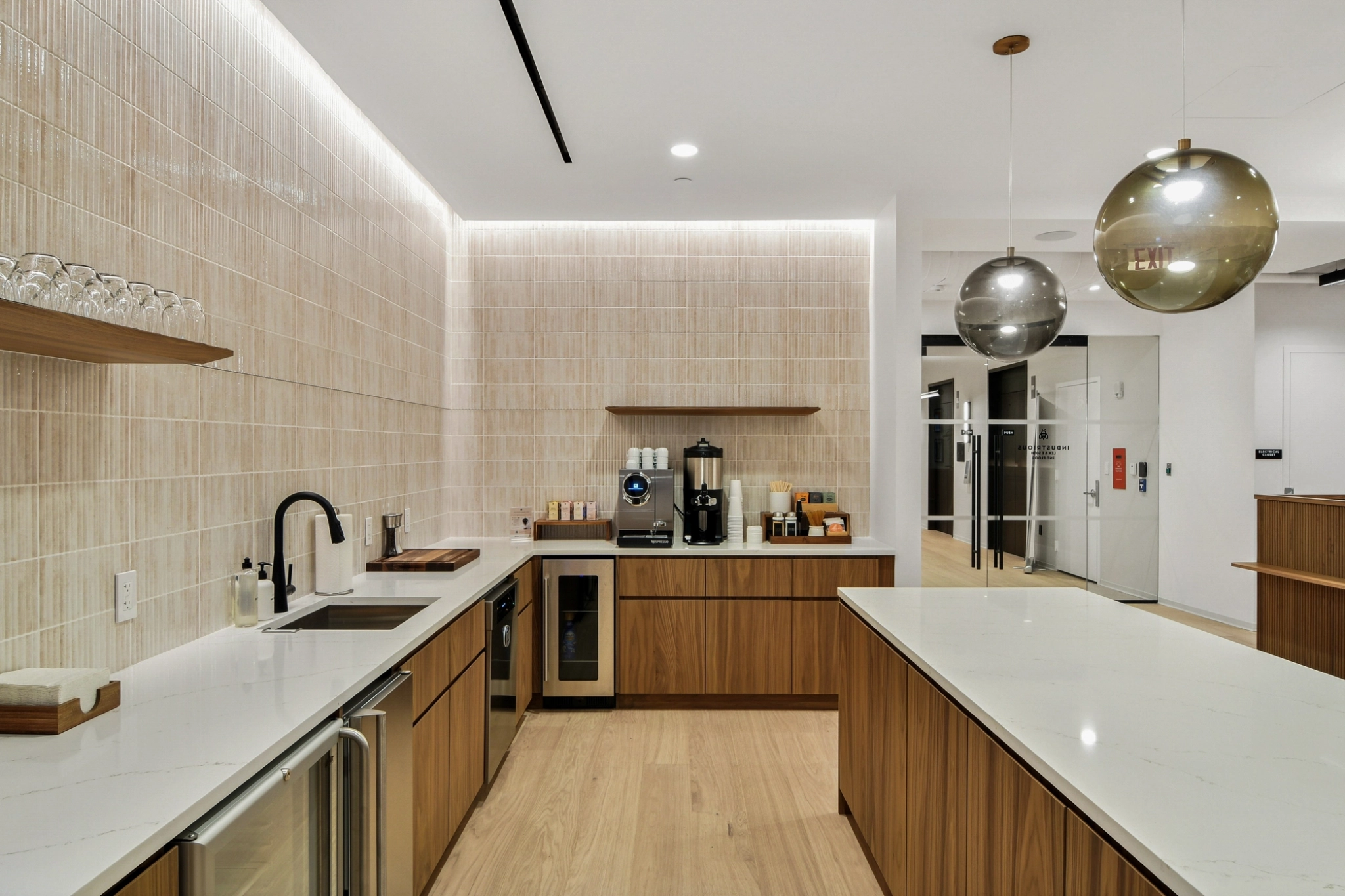 Cuisine moderne avec placards en bois, plans de travail blancs, appareils électroménagers intégrés, machine à café, étagères en verre et deux lampes suspendues à globe, parfaite pour un espace de coworking ou un bureau élégant à New York. Les murs en carrelage clair et le sol en bois complètent l'ensemble.