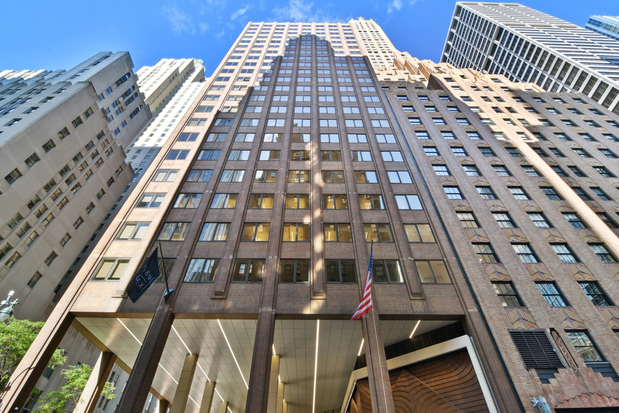 Un grand immeuble de bureaux à la façade de briques brunes, vu d'un angle bas, propose des espaces de coworking et des salles de réunion modernes, avec des drapeaux américains à l'entrée et d'autres gratte-ciel à proximité.