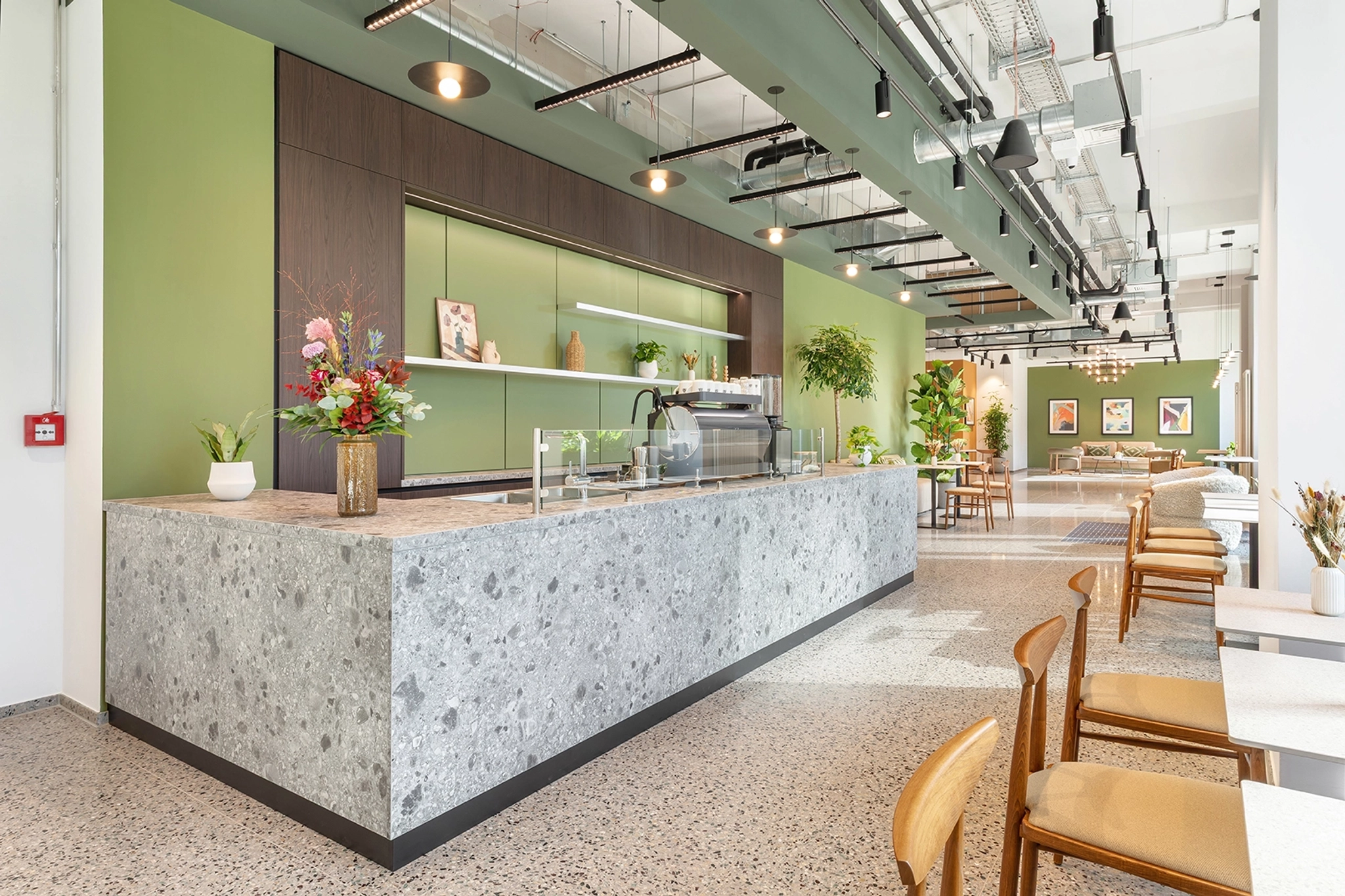 Modern Berlin cafe with a large stone counter, wooden chairs, green walls, pendant lighting, and potted plants; seating area and framed art visible in the background—perfect for an office break or informal meeting room ambiance.