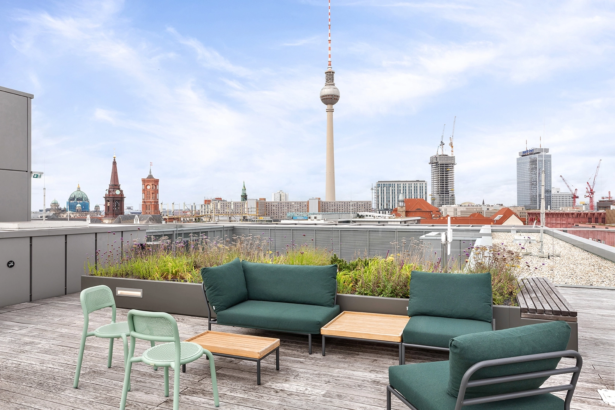 Rooftop terrace with green cushioned seating and wooden tables, offering city views including the Berlin TV Tower—perfect for a relaxing workspace or coworking spot under a partly cloudy sky.