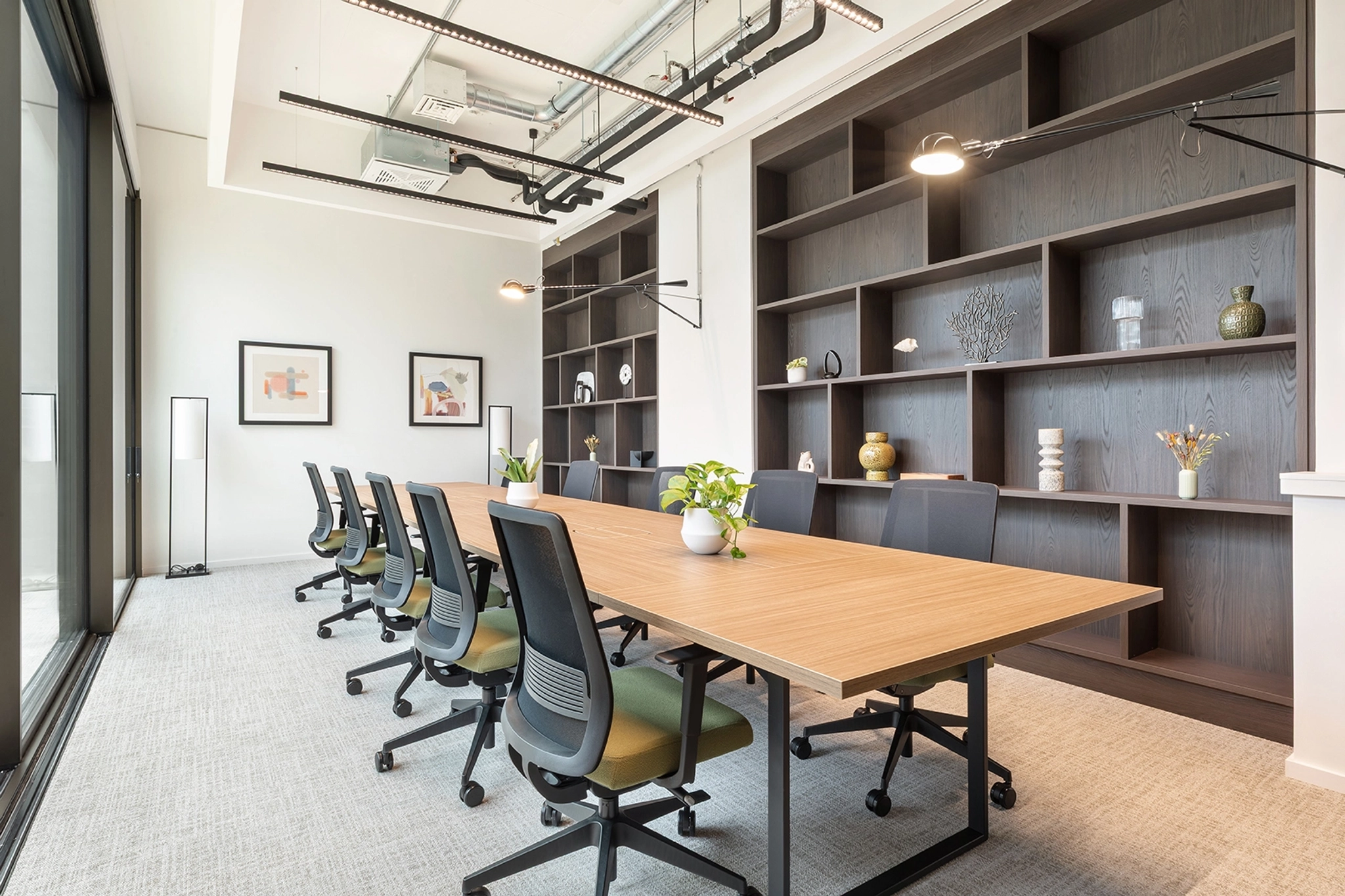 Modern coworking conference room with a long wooden table, office chairs, open shelving, potted plants, framed art, and large windows letting in natural light—perfect for an inspiring workspace.