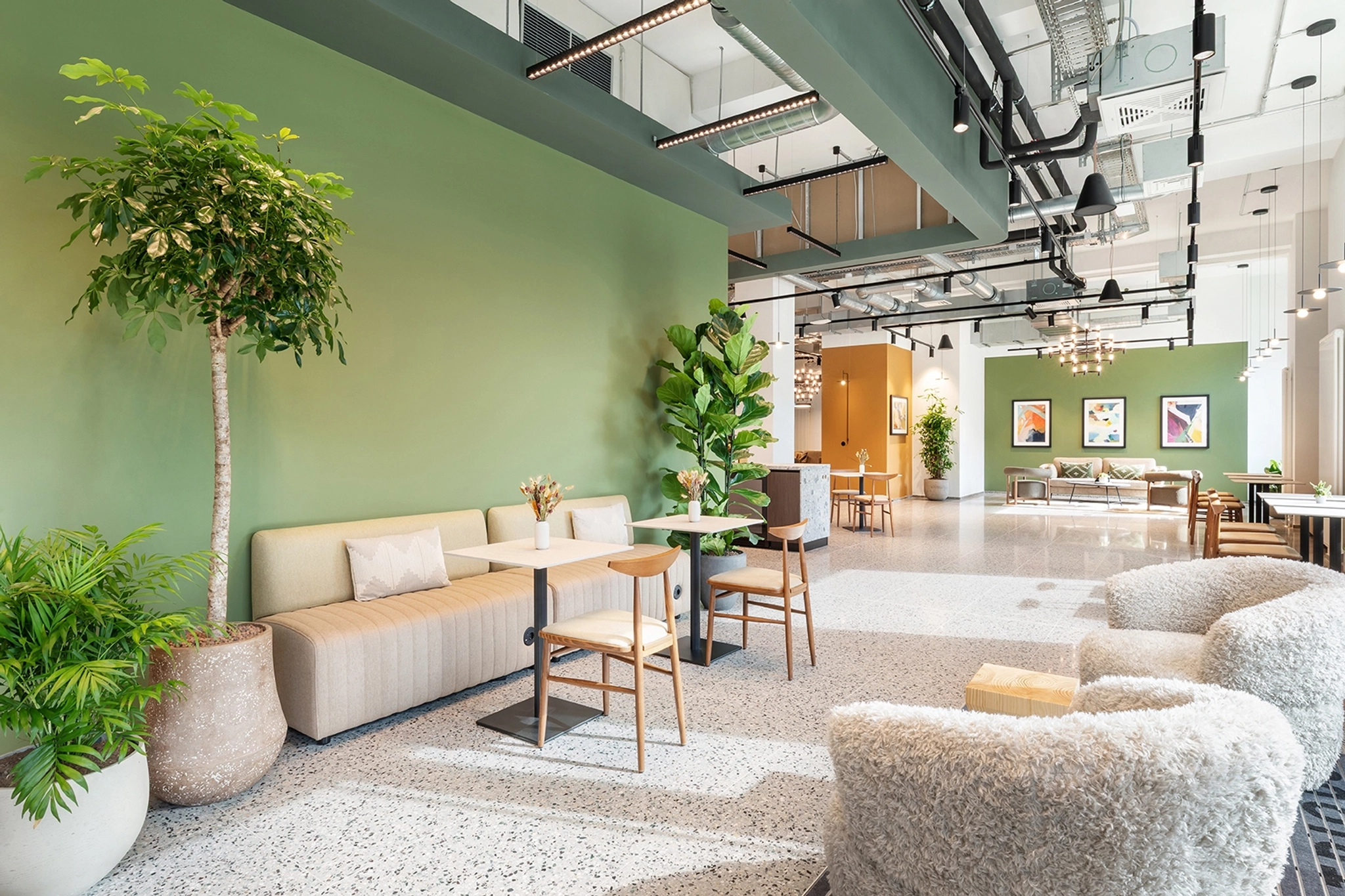 Modern lounge area in a Berlin office with green walls, potted plants, beige sofa, wooden chairs, small tables, and soft lighting in a spacious, contemporary interior—perfect for casual meetings or relaxing outside the meeting room.