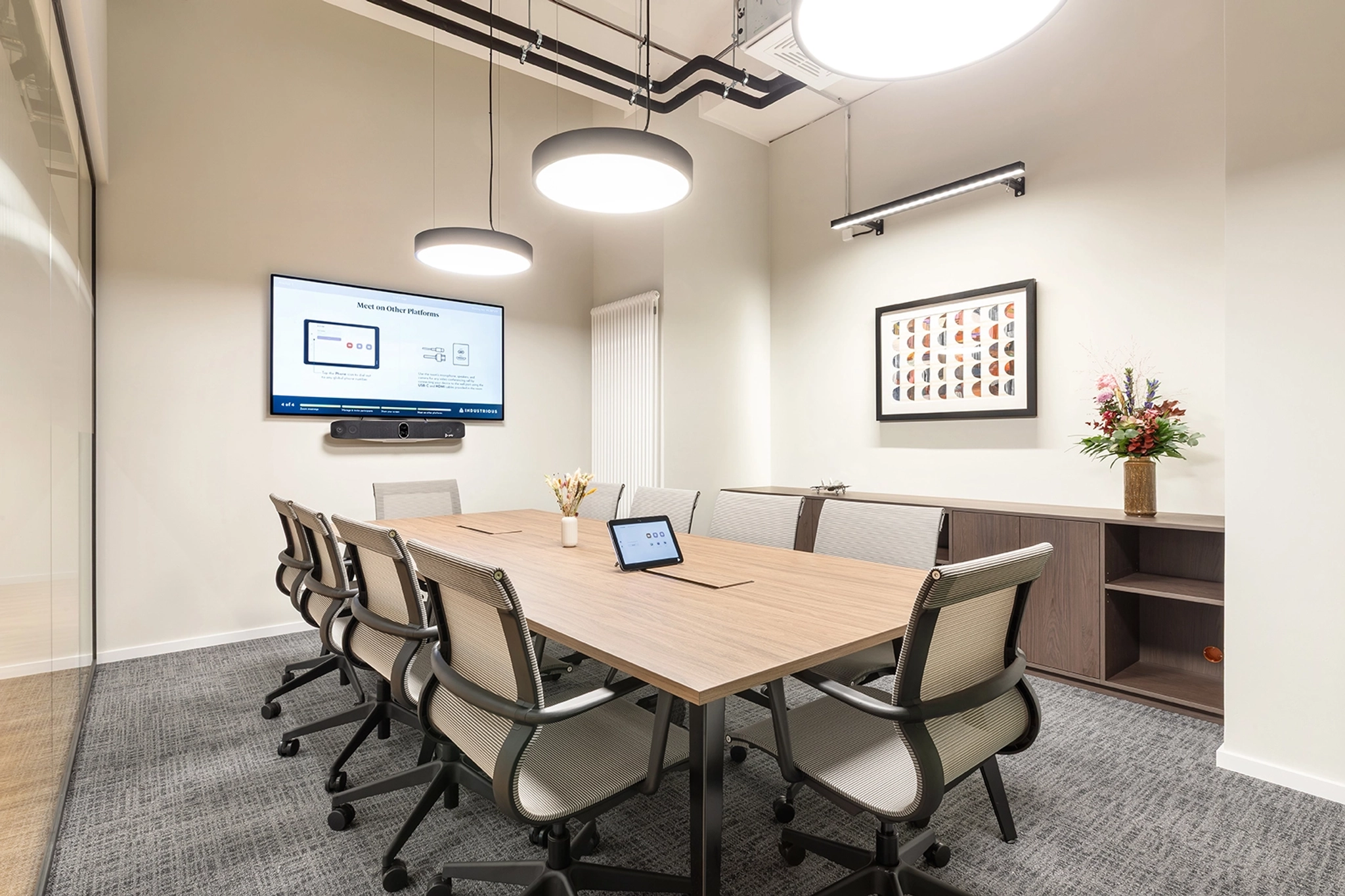 A modern meeting room with a long table, eight chairs, a wall-mounted screen displaying a presentation, overhead lights, and a side cabinet with a vase of flowers and framed art—perfect for any coworking office.