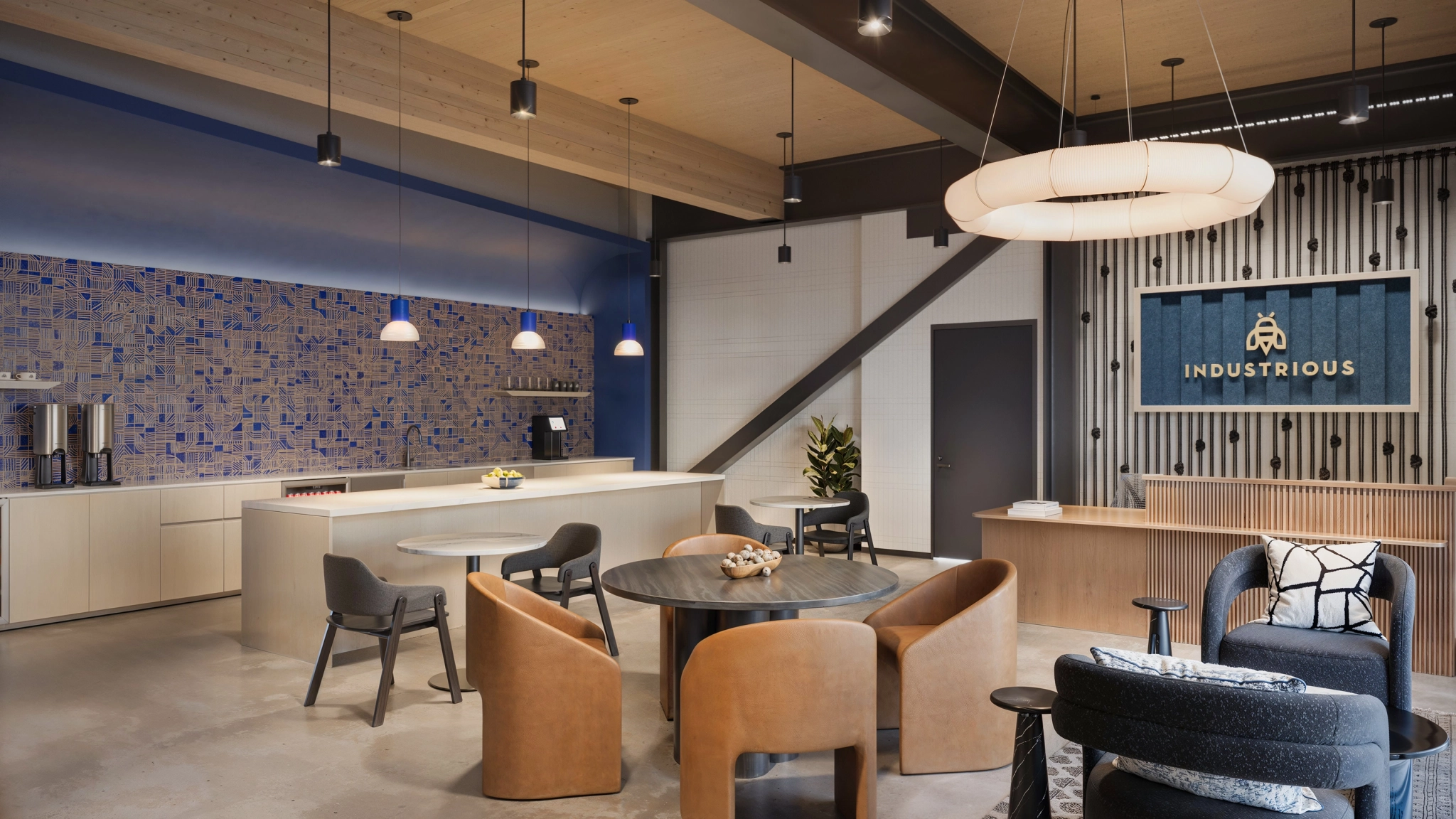 Modern office lounge with round tables, leather chairs, a coffee station, pendant lights, and an "Industrious" sign on the wall. A blue accent wall and various workspace seating options are visible.