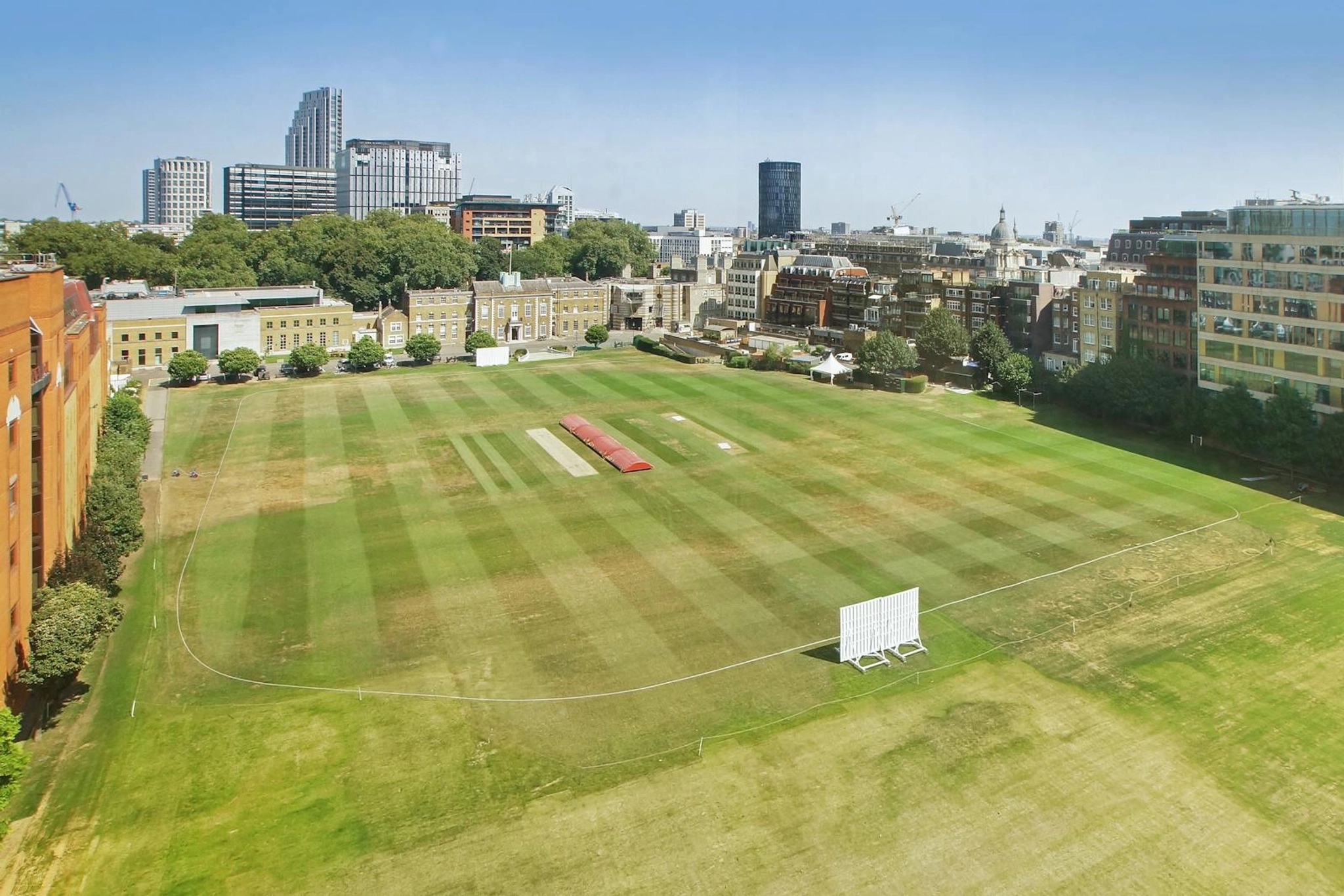 Aerial view of a cricket field with boundary lines, wicket strips, and city office buildings in the background on a clear day.