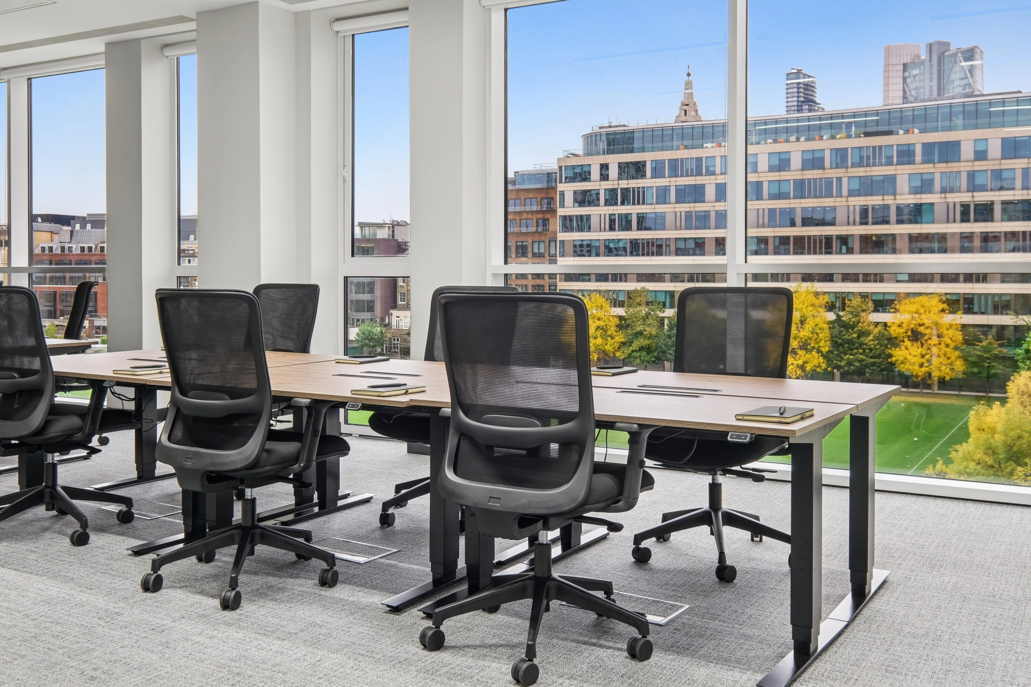 Modern London office space with empty black chairs and desks, facing large windows that overlook city buildings and a leafy park. Daylight streams into the workspace, creating a bright and inviting atmosphere.