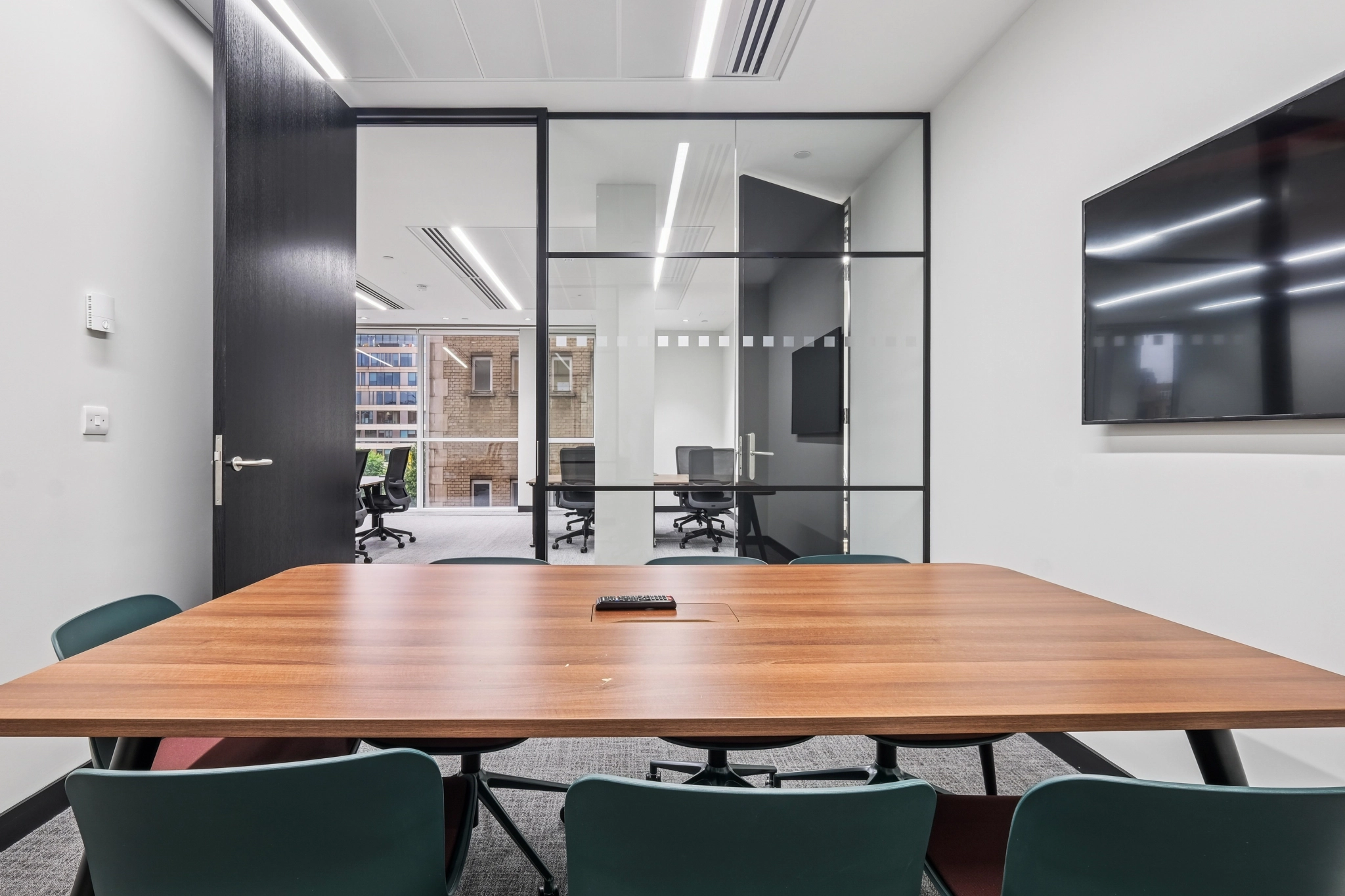 Une salle de réunion moderne avec une table en bois, des chaises vertes, une télévision murale et des portes vitrées qui s'ouvrent sur un espace de travail adjacent rempli de fenêtres.