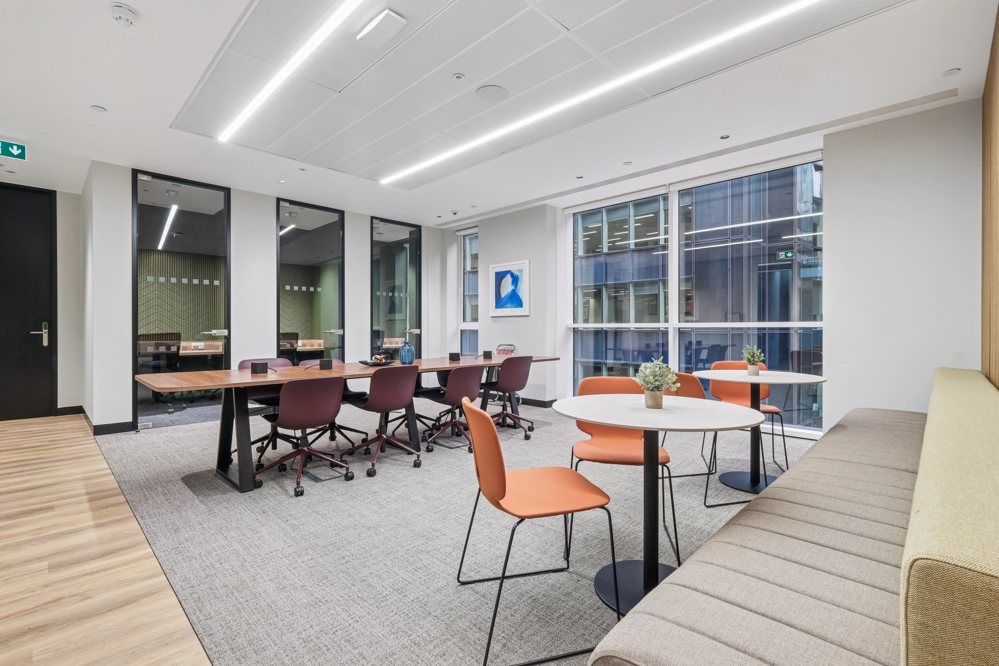 Bureau moderne à Londres comprenant une salle de réunion spacieuse avec une longue table, des chaises violettes, deux tables rondes avec des chaises orange, un banc beige, de grandes fenêtres et des plantes d'intérieur luxuriantes.