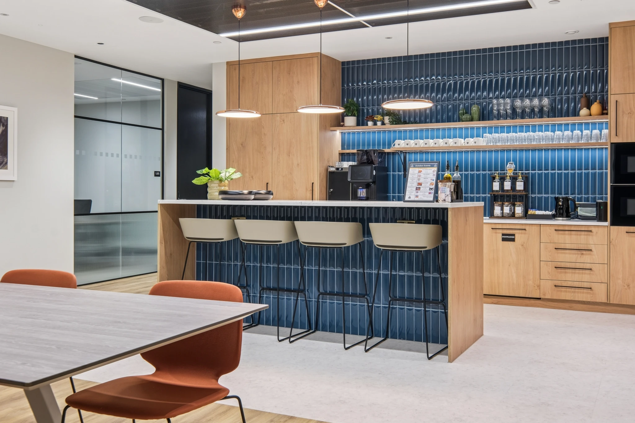 Modern office kitchen in London with wood cabinets, blue tile backsplash, a counter with four stools, coffee machines, shelves with cups, and a table with orange chairs creates a vibrant workspace atmosphere.