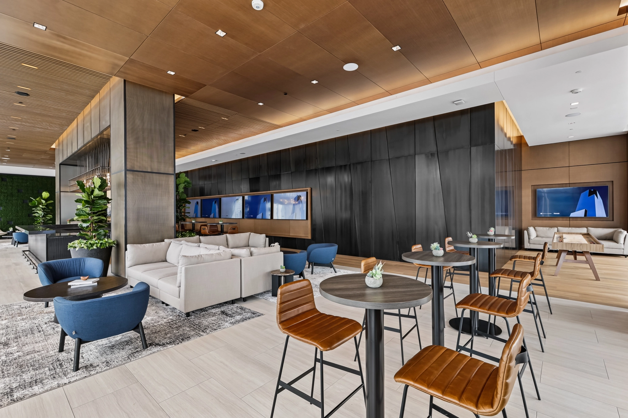 Modern office lounge area with blue chairs, sofas, tall tables with brown stools, potted plants, light wood flooring, and large wall-mounted screens in a well-lit space—perfect for informal meetings in Nashville.