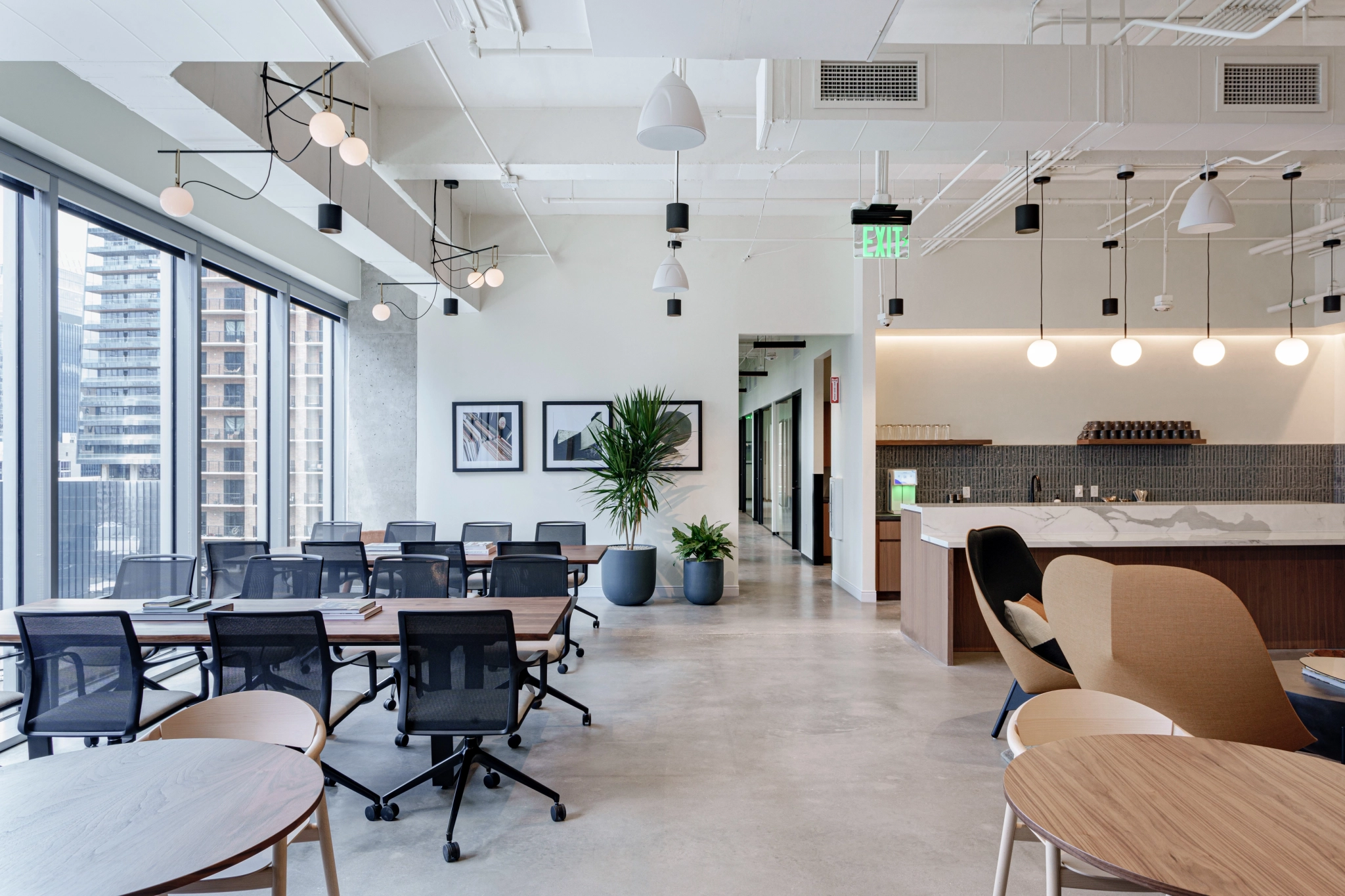 Espace de bureau moderne à New York avec de grandes fenêtres, plusieurs bureaux et chaises, un coin salon, des plantes d'intérieur et une kitchenette à l'arrière-plan.