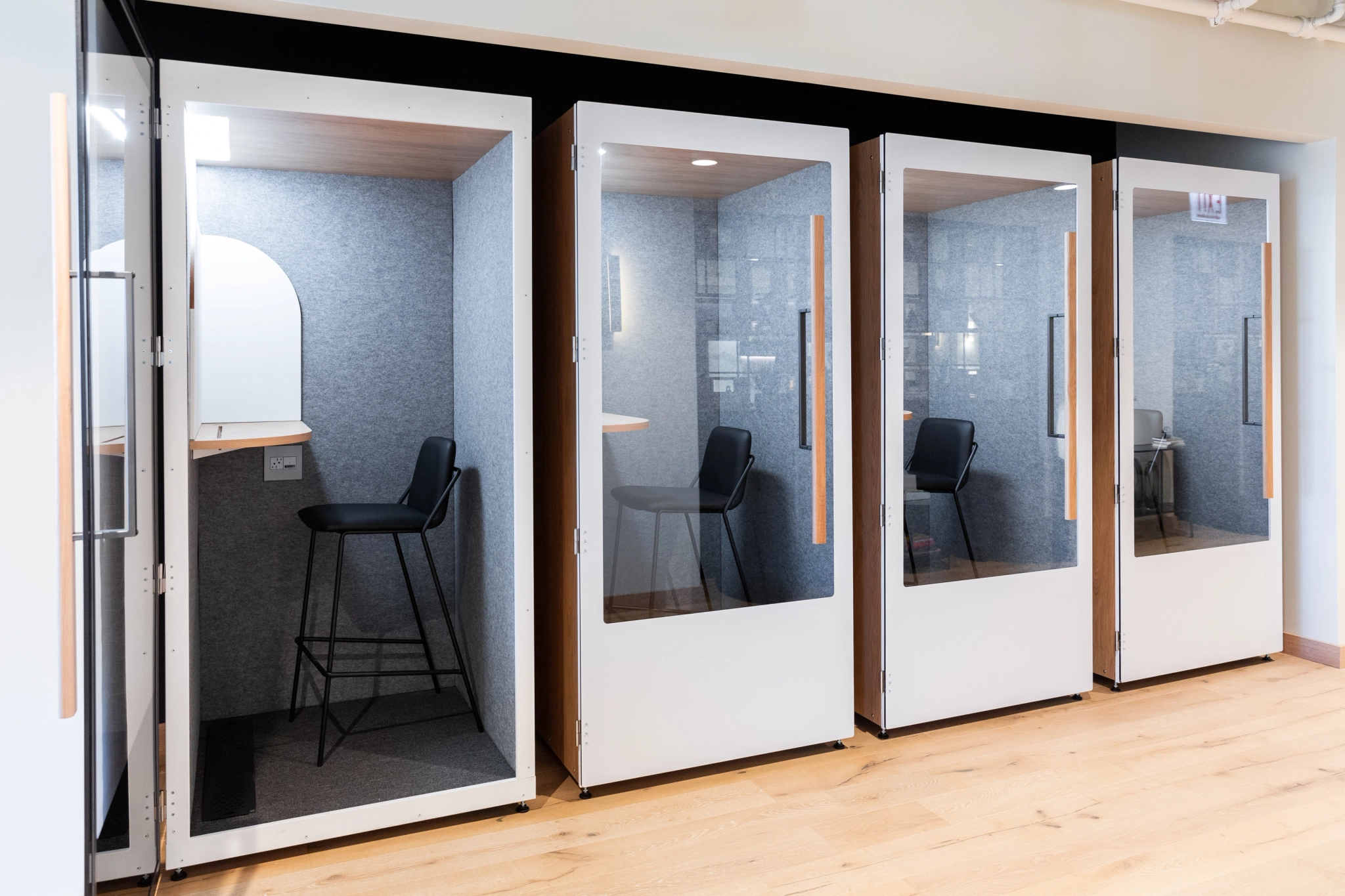 Four modern soundproof phone booths with glass doors, stools, desk surfaces, and light wood finishes are lined up in this coworking office space with matching light wood flooring.