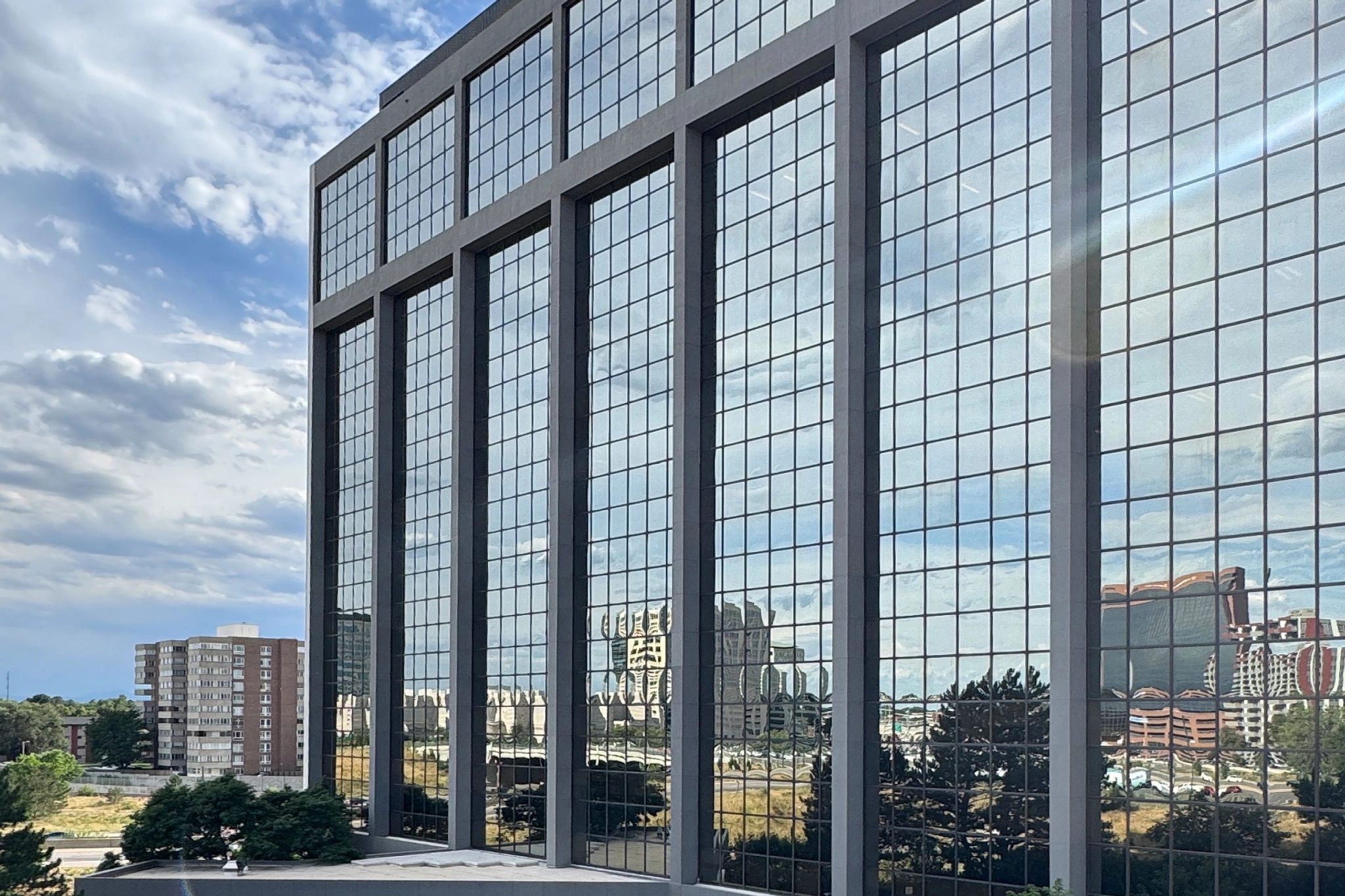 A tall glass office building reflects the sky, clouds, and nearby structures on its large window panels, with trees and other buildings visible in the background—perfect for a modern workspace environment.