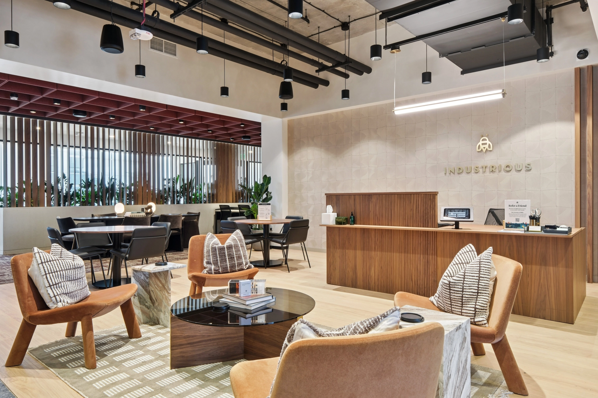 Moderne kantoorlounge met houten meubilair, kussens, een glazen salontafel, een open werkruimte en een receptie met een "Industrious"-bordje - perfect voor coworking of informele vergaderingen.