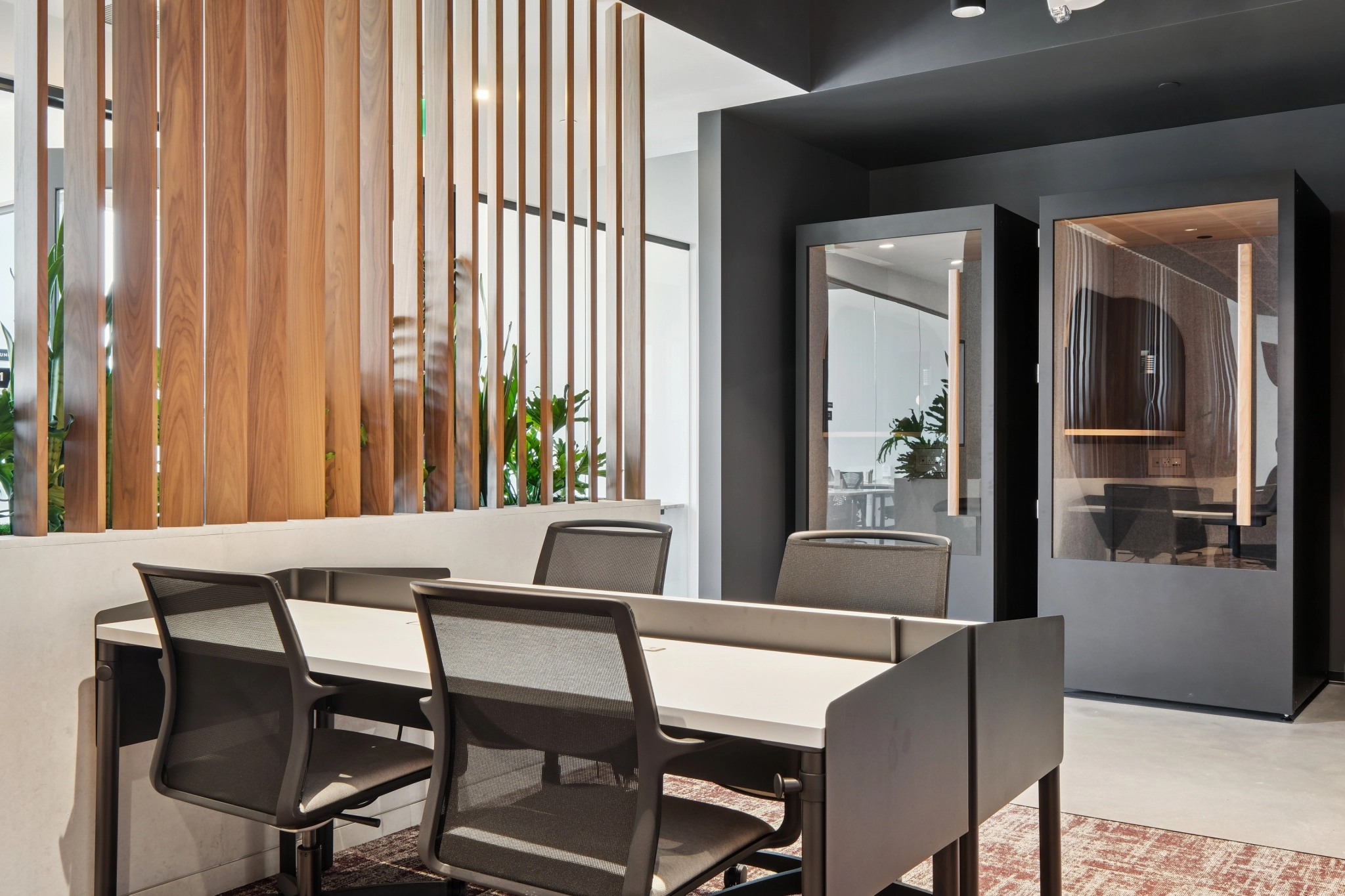 Moderner Coworking-Space mit einem rechteckigen Tisch, vier Gitterstühlen, einer Trennwand aus Holzlatten und einer geschlossenen Telefonzelle im Hintergrund - perfekt für New Yorker Berufstätige.