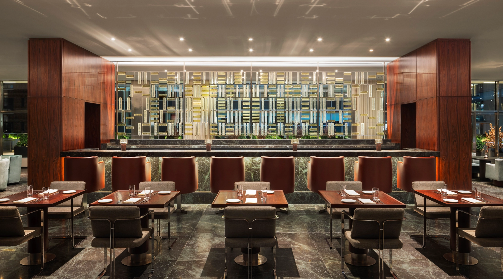 Intérieur de restaurant moderne avec sols en marbre foncé, tables et chaises en bois, et écran métallique décoratif derrière le comptoir du bar. Parfait pour des réunions décontractées ou pour travailler dans un espace de travail élégant.