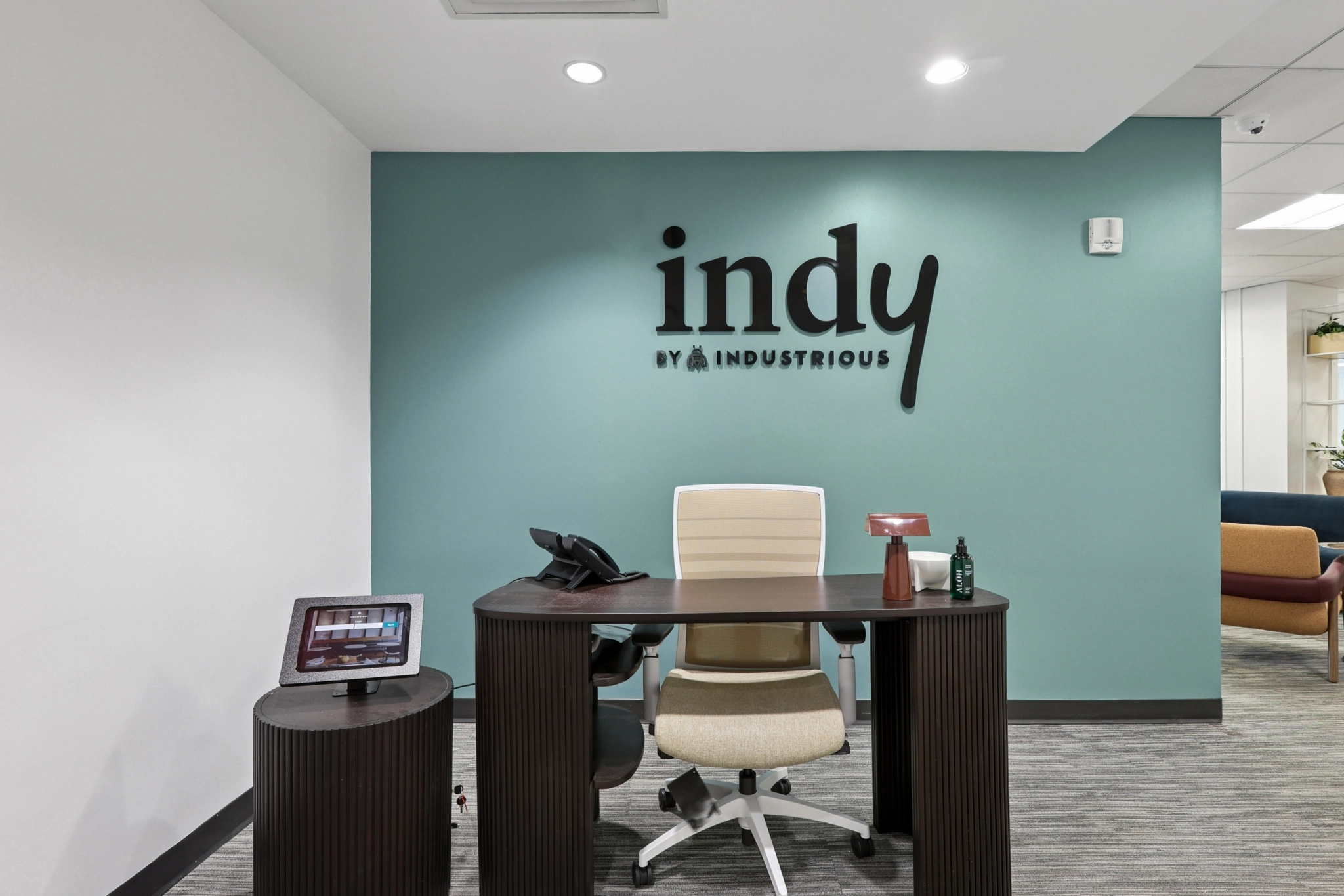 Reception desk with a chair, phone, tablet, and office supplies in front of a blue-green wall with "indy BY INDUSTRIOUS" signage, creating a modern coworking workspace setting.