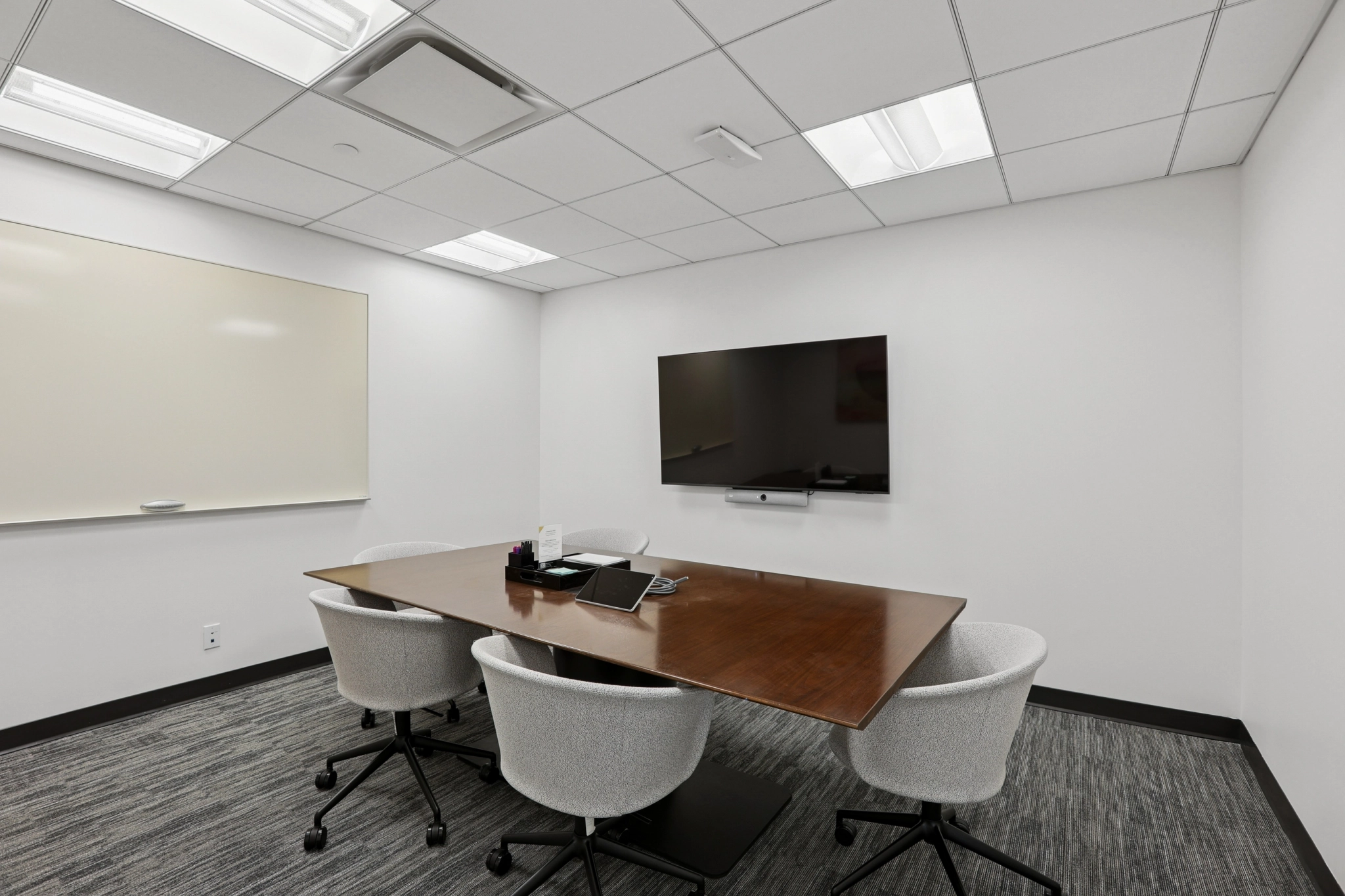 A small coworking conference room in New York, featuring a wooden table, four chairs, a wall-mounted TV, a whiteboard, and a tablet on the table—perfect for your next office meeting.