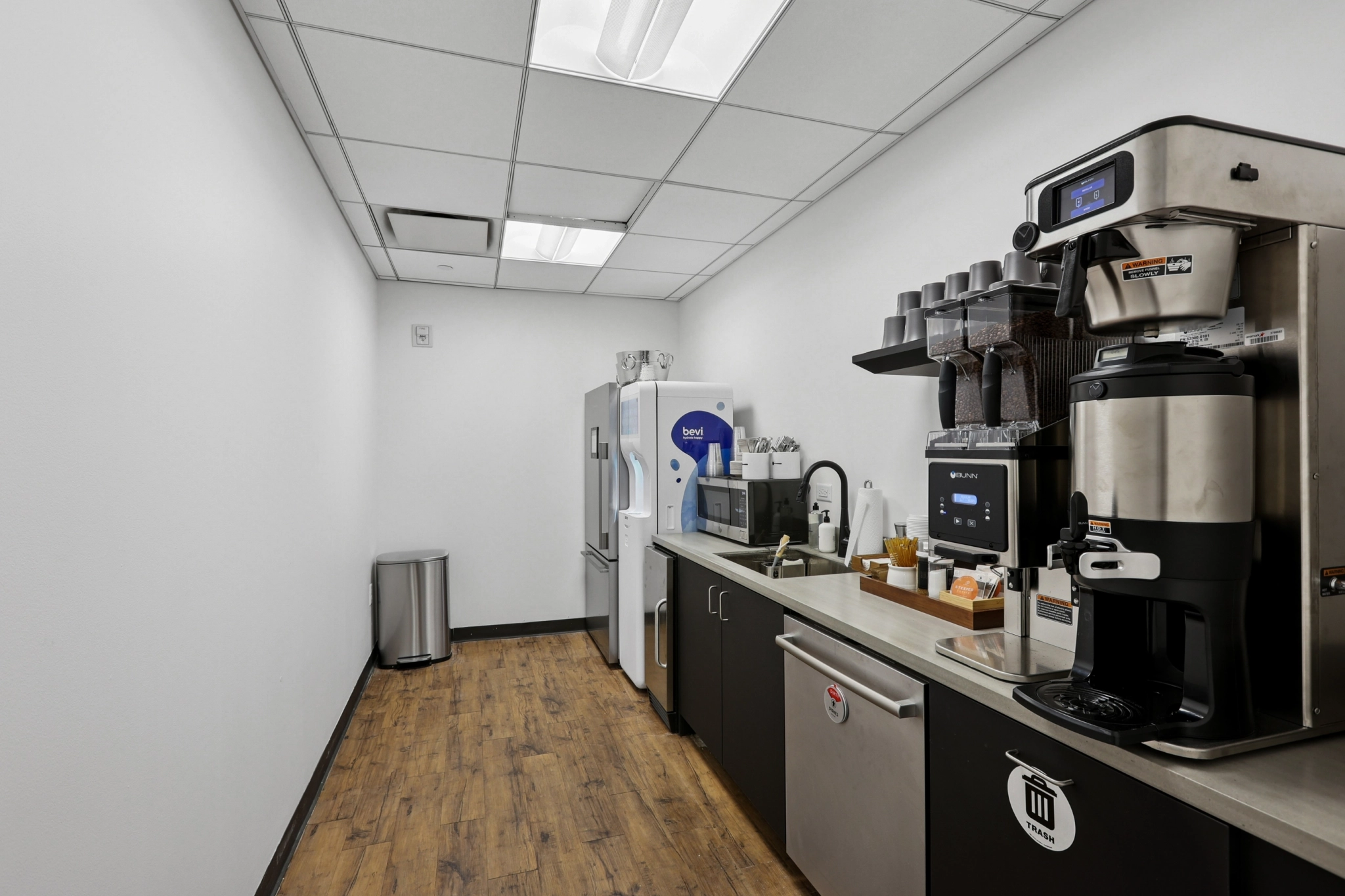 Small break room in an office with a coffee machine, countertop with supplies, refrigerator, microwave, sink, and a trash can on wood flooring under fluorescent lights—perfect for coworking or a quick pause between meeting room sessions.