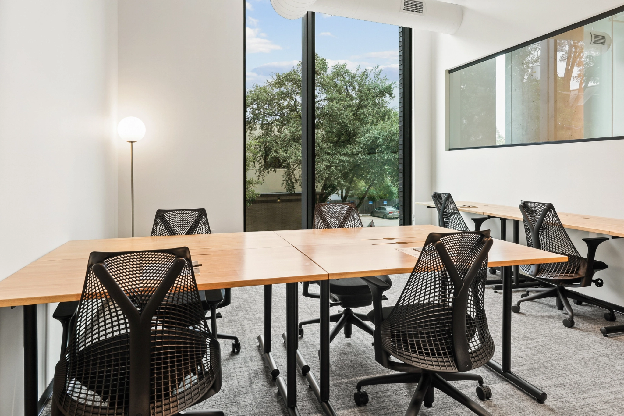Moderner Arbeitsbereich in Boston mit sechs schwarzen Netzstühlen um helle Holzschreibtische, grauem Teppichboden, einem großen Fenster mit Blick auf die Bäume, einer Glastrennwand und einer Stehlampe - perfekt für ein Besprechungszimmer.