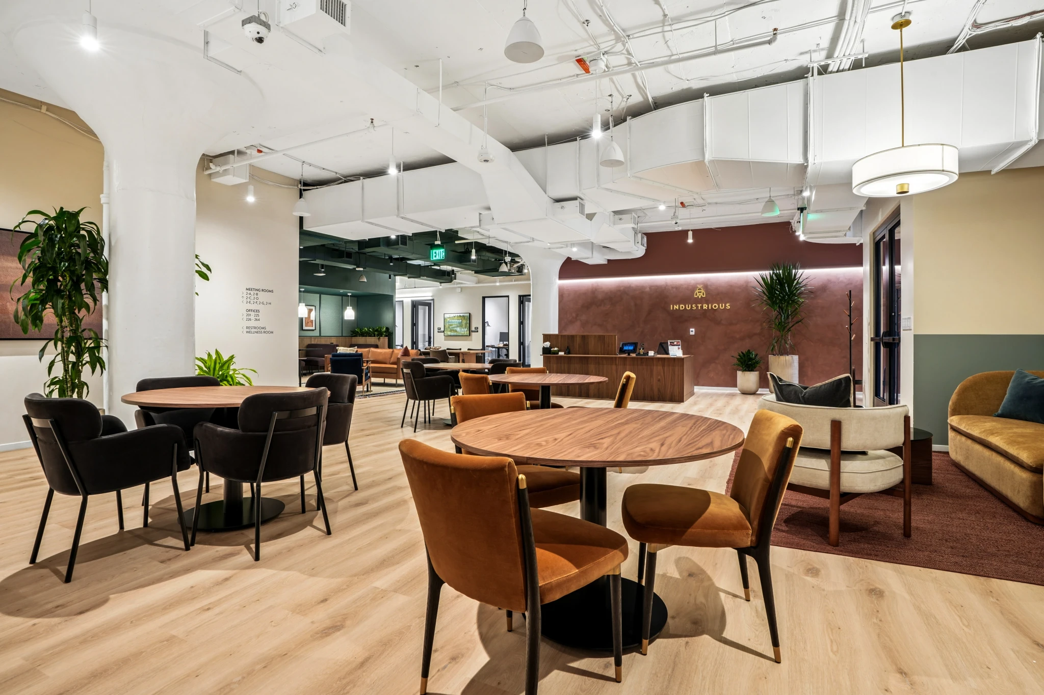 Modern Boston office lounge with round wooden tables, mixed chairs, lush plants, light wood flooring, and a coworking reception desk in the background under bright ceiling lights.
