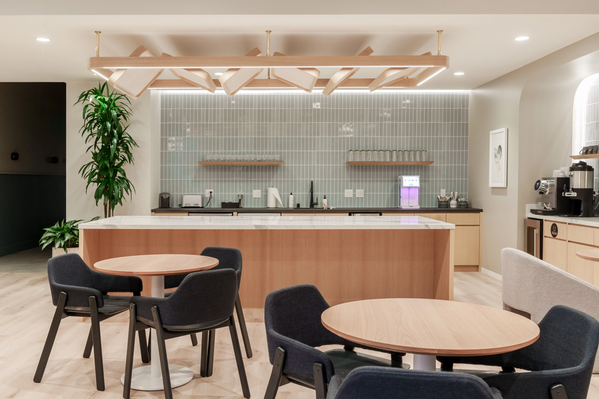 Modern office kitchen and break area features round tables and chairs, a large island, tiled backsplash, coffee machine, potted plant—perfect for relaxing or collaborating in a contemporary coworking workspace.