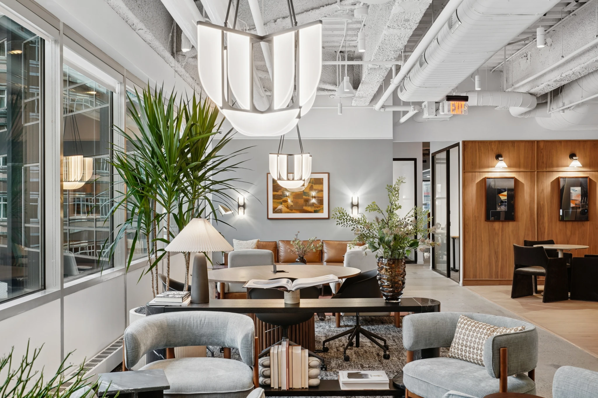 Una oficina moderna en Nueva York con grandes ventanales, mobiliario contemporáneo, lámparas colgantes, plantas de interior y detalles en madera. La zona de descanso diáfana y las pequeñas salas de reuniones que se ven al fondo crean un entorno de trabajo acogedor.