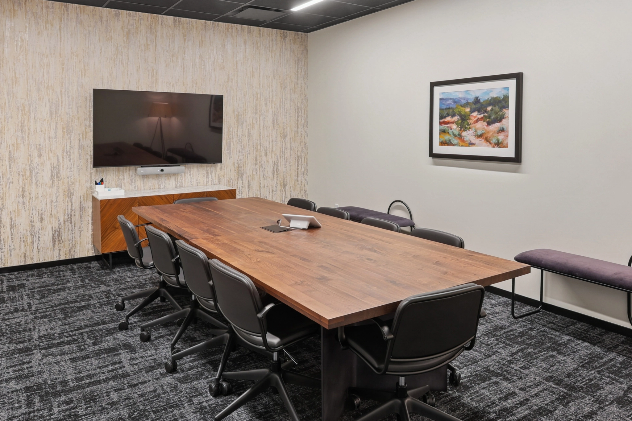 A modern coworking conference room with a wooden table, black chairs, a wall-mounted TV, a framed landscape painting, and a bench along the right wall.