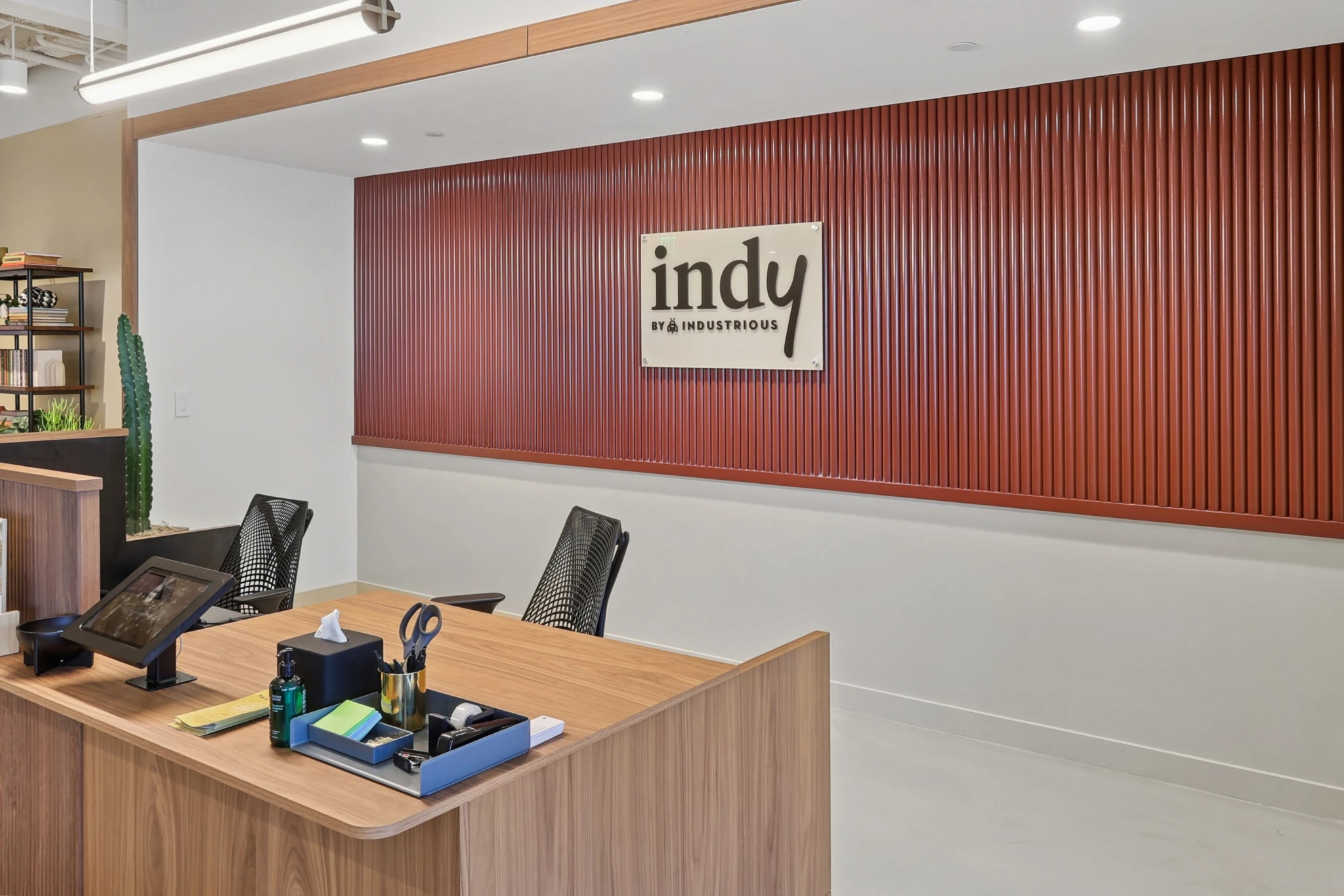 A modern office reception area with a wooden desk, two chairs, office supplies, and a red accent wall displaying a sign that reads "indy by Industrious" welcomes you to this vibrant workspace.
